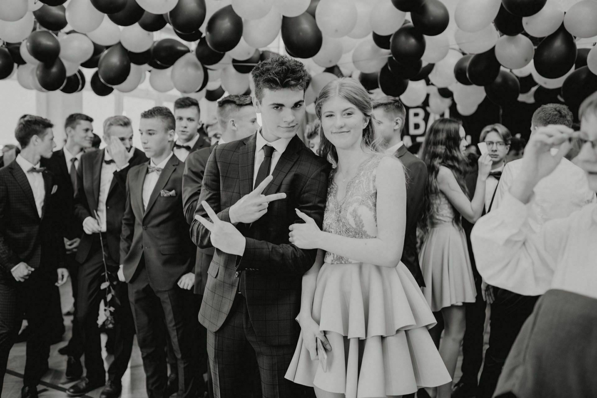 The black and white photo shows a group of teenagers at a party under a ceiling decorated with black and white balloons. Two teenagers in the foreground, dressed formally, pose for this classic event photo, while a boy shows a peace sign and both smile for the camera. 