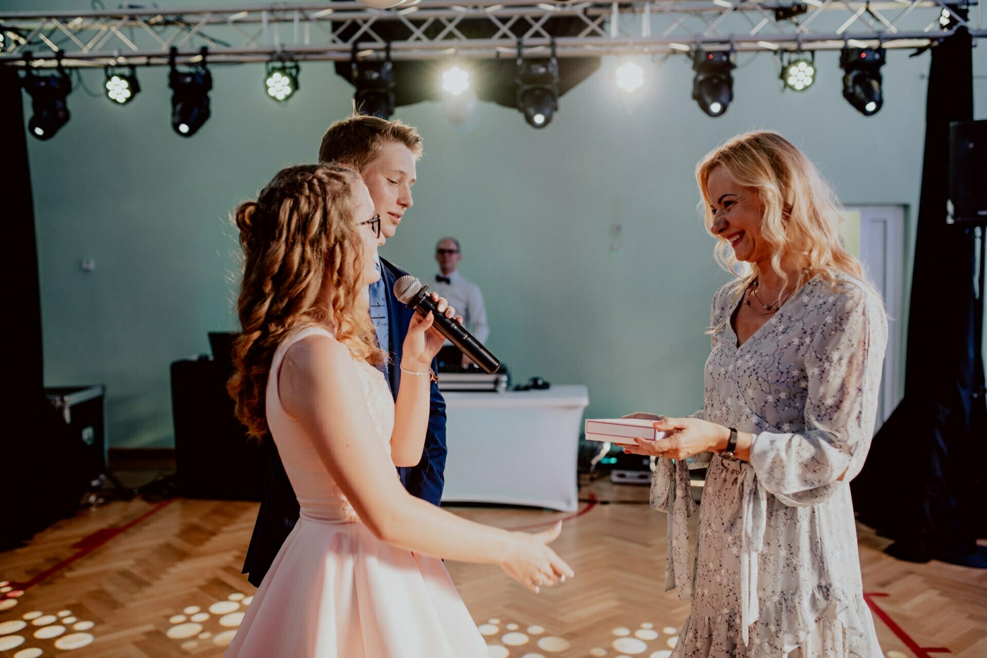 A young woman in a pink dress speaks into a microphone and gestures toward a smiling woman in a light blue dress who is holding a book. Next to the woman with the microphone stands a young man in a navy blue suit. Bright stage lights illuminate the scene, perfect for event photography.  