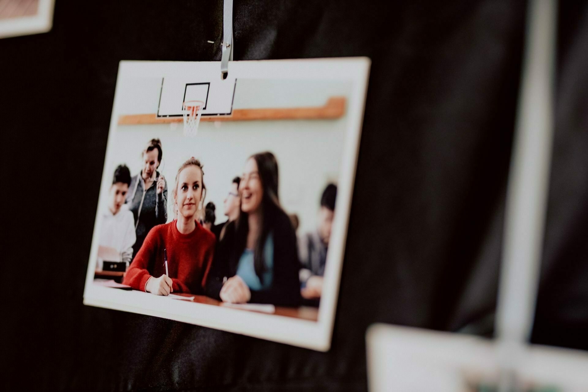 The event photo shows three students in a classroom: one in a red top focuses on writing, while the two next to her smile. This evocative event photo is cut out and hung against a dark background. 