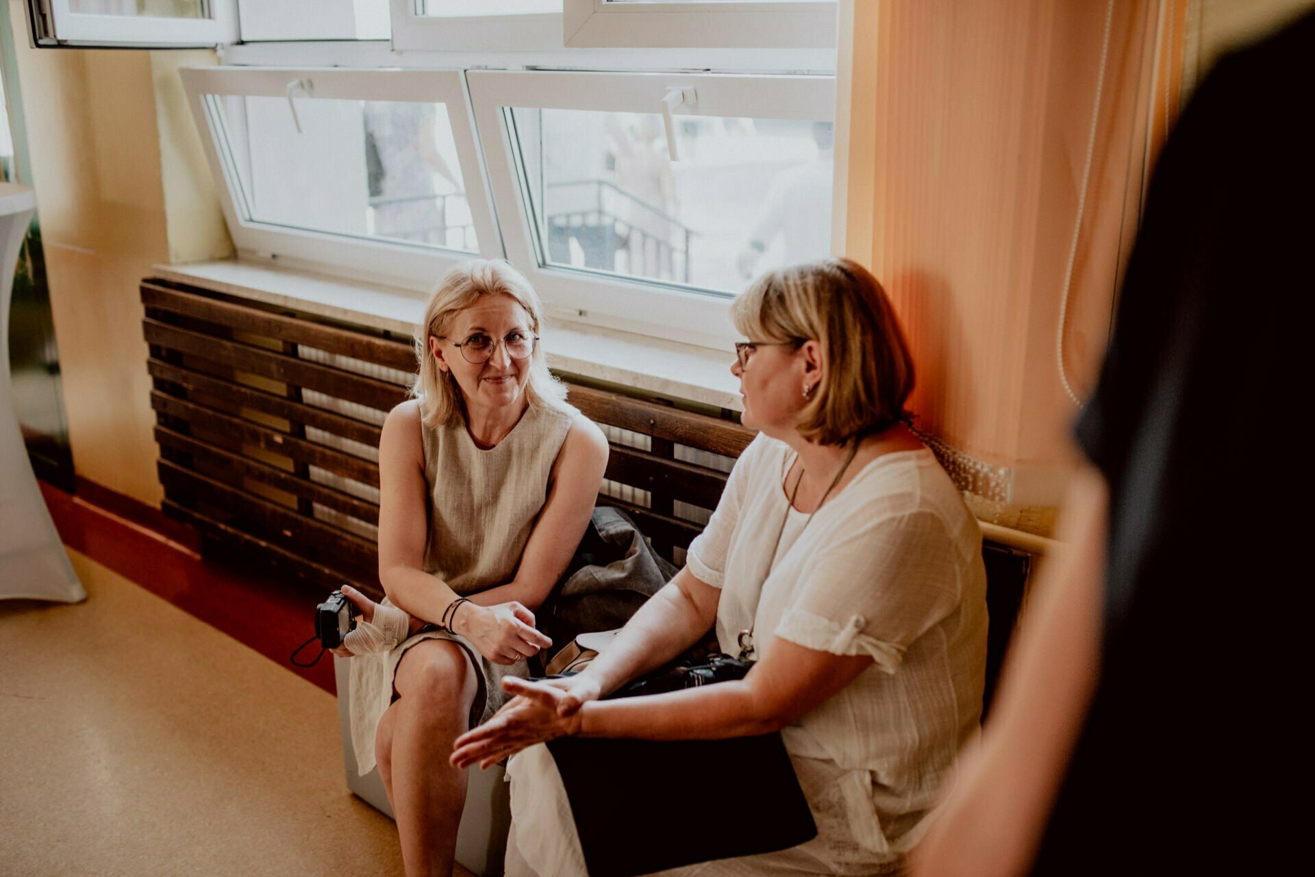 Two women are sitting on a bench in a room and talking to each other. One of the women has blond hair, wears glasses and is dressed in a light beige outfit. The other woman, also blonde and wearing glasses, is dressed in white. The windows are in the background, capturing the perfect moment for event photography.   