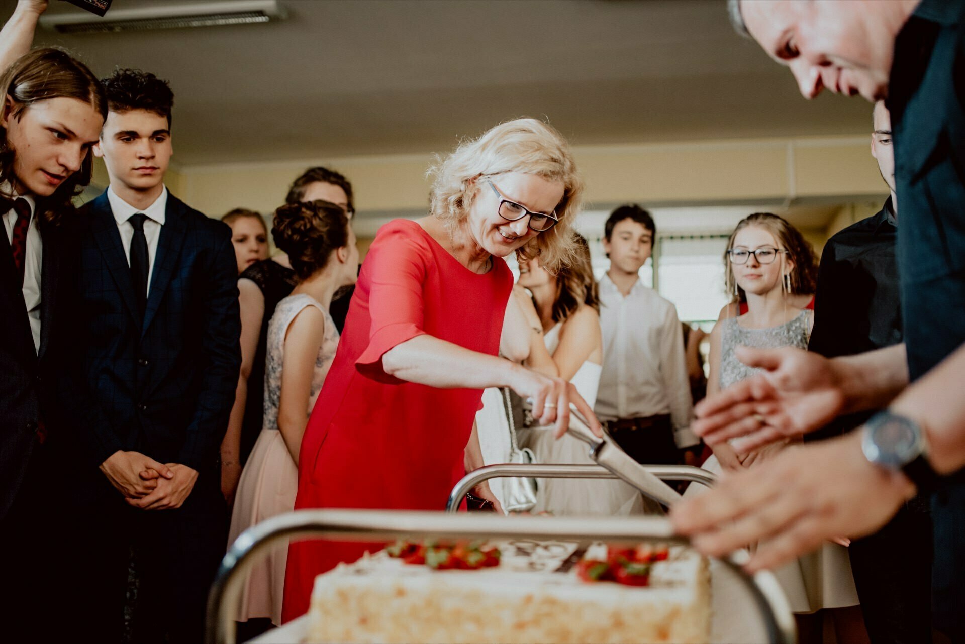 A woman in a red dress uses a large knife to cut a cake on a metal cart. She is surrounded by a group of people dressed formally. Some are clapping, others are eagerly watching the event in the well-lit room, which was beautifully captured by event photographer Warsaw.  