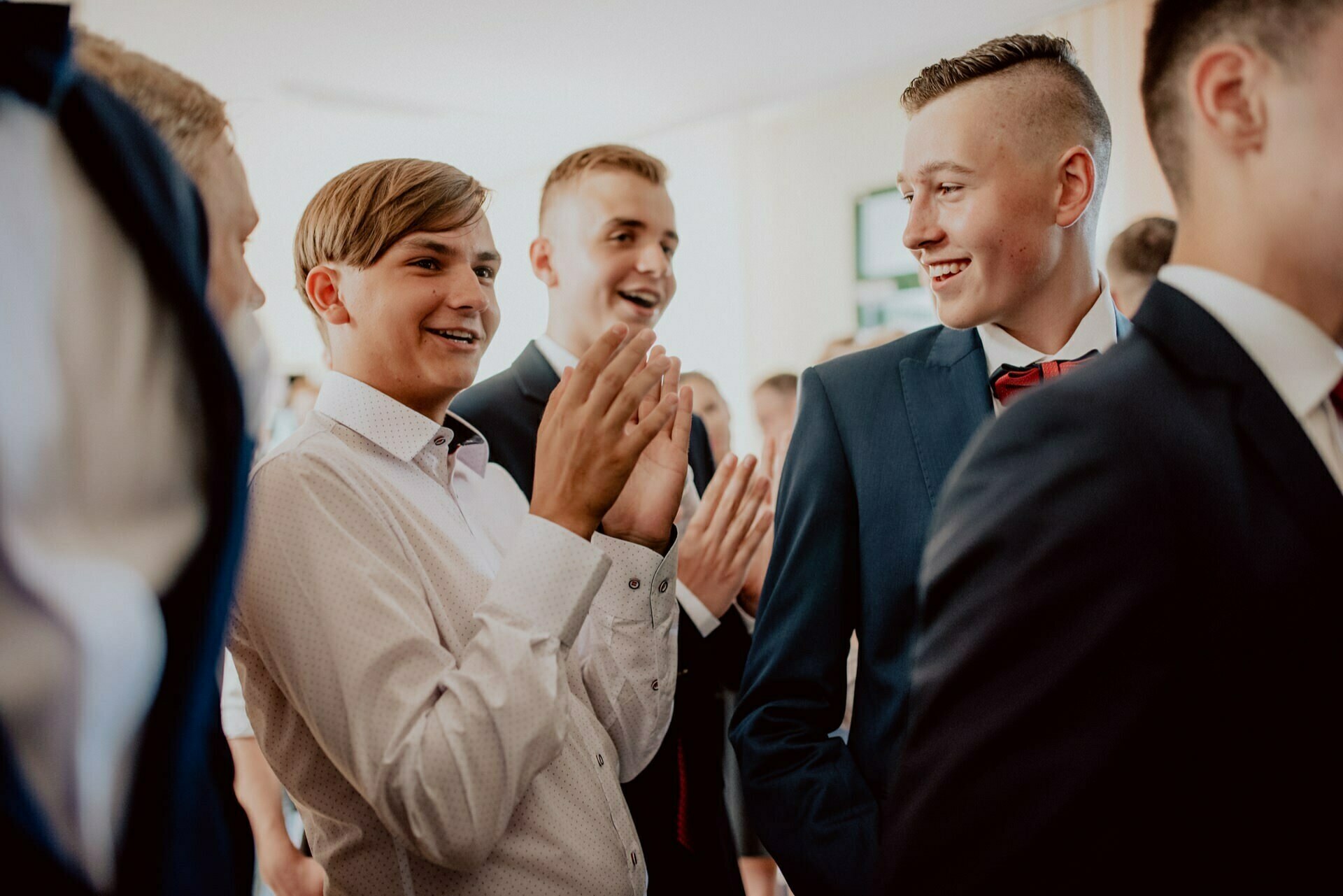 A group of young men dressed in formal attire smile and laugh together in the room. One man in a white shirt claps, while others in suits look on cheerfully. The atmosphere seems lively and joyful - perfectly captured by event photography.  