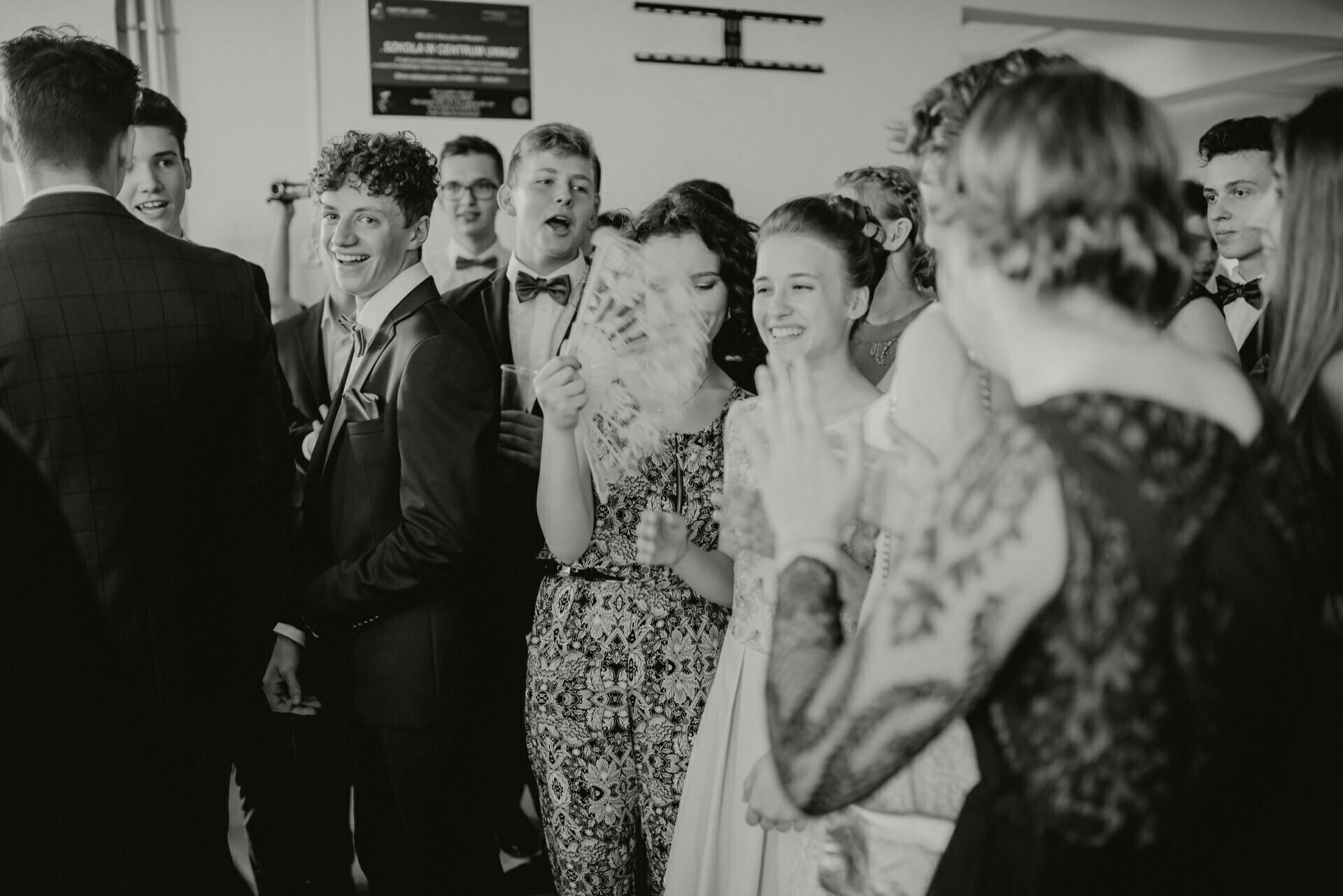 A black and white photo of a group of people dressed in formal attire at a lively social event. They appear to be having a good time, some smiling and some clapping. One person is holding a decorative fan. This event photography perfectly captures the festive mood.   