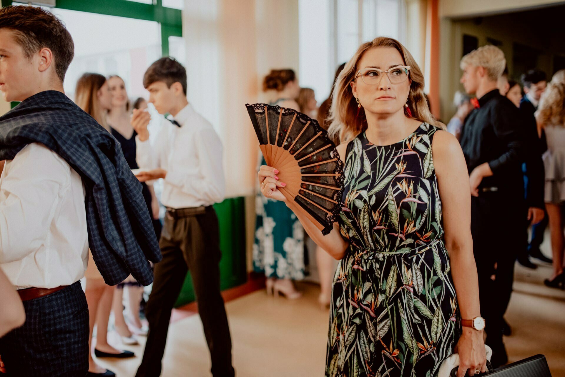 A woman in a flowery dress and glasses holds an ornate fan in a crowded room. She looks to the side while people around her carry on a conversation. The scenery appears to be a formal event or gathering, beautifully captured by event photographer Warsaw.  