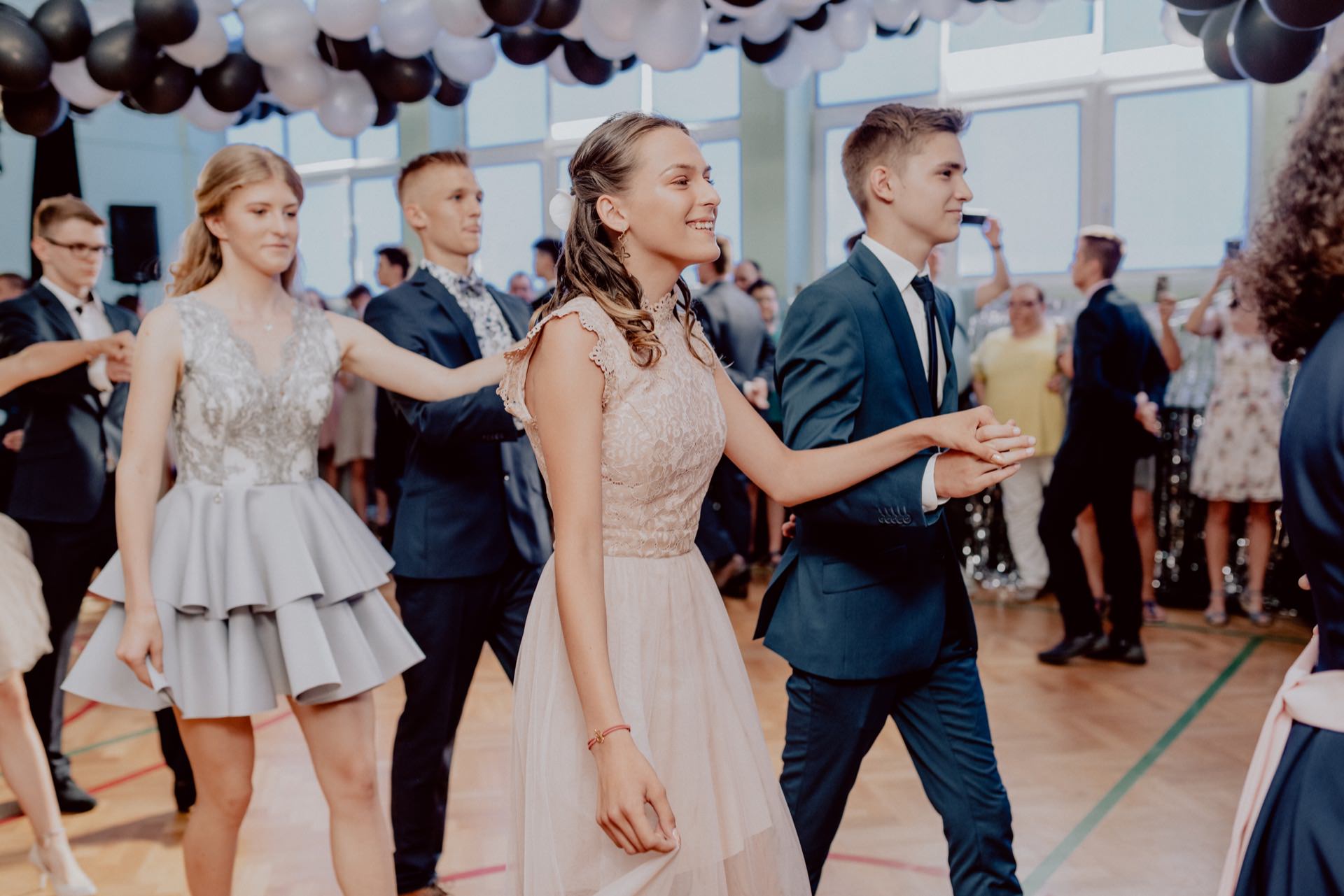 A group of young people dressed in formal attire are dancing in pairs in a decorated room, probably at a prom or wedding reception. Balloon decorations float above them, while other guests watch them from the background. This scene could be perfectly captured by an event photography specialist, providing a great photo report of the events.  