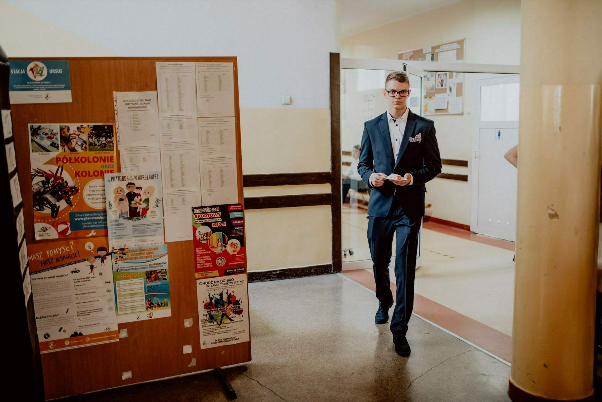 A man in a blue suit is standing in a hallway next to a bulletin board filled with various posters and announcements, beautifully captured in this event photograph. He holds a small object in his hands and seems to be walking or taking a step forward. The space behind him is bright and lightly furnished.  