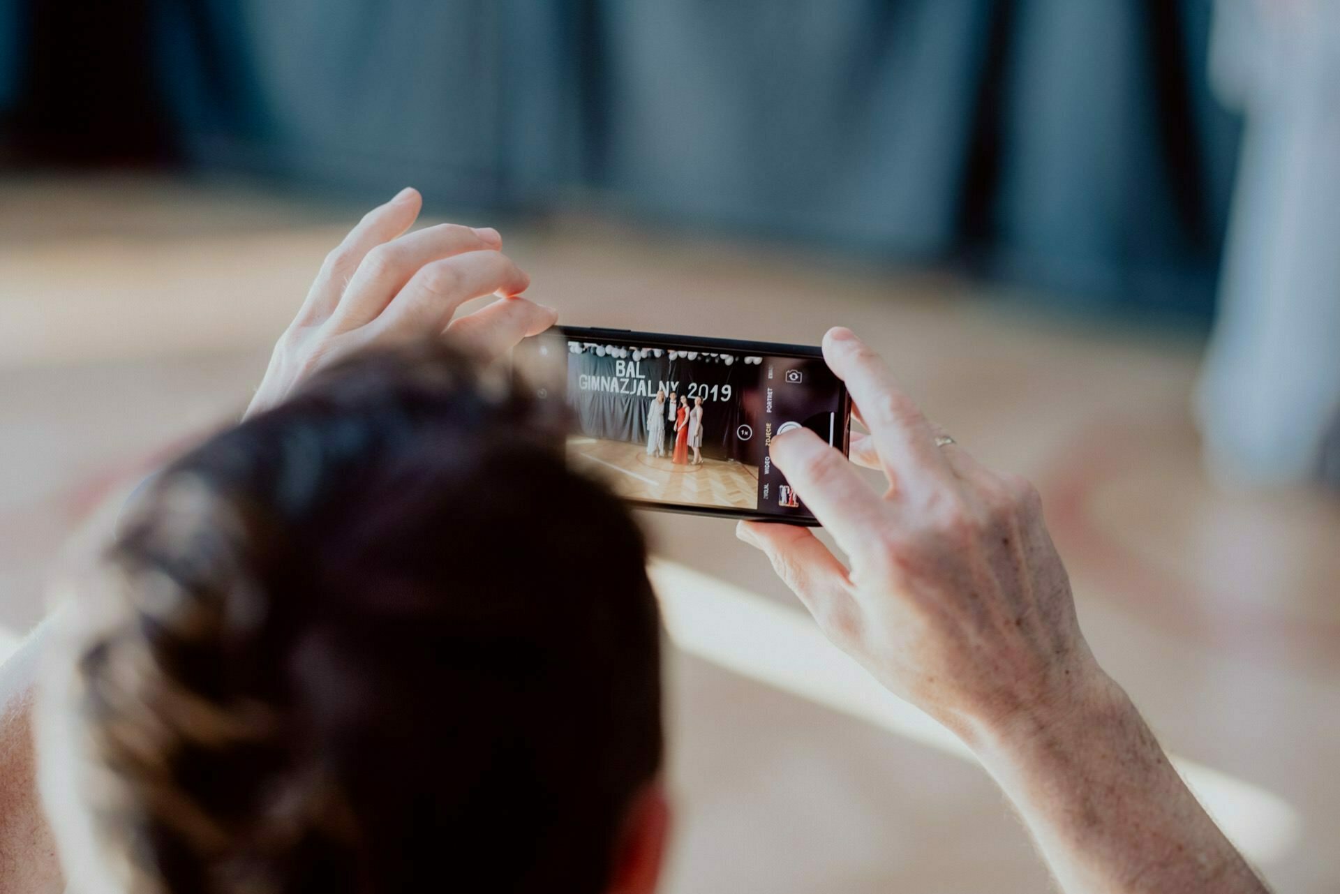 A person holding a smartphone horizontally and taking a photo or video of a dance event with a banner reading "BALI CINNAMONZALIY 2019". The person's head is slightly out of focus, and the phone displays the scene in greater detail - ideal for any event photographer Warsaw presenting a photo report on events. 