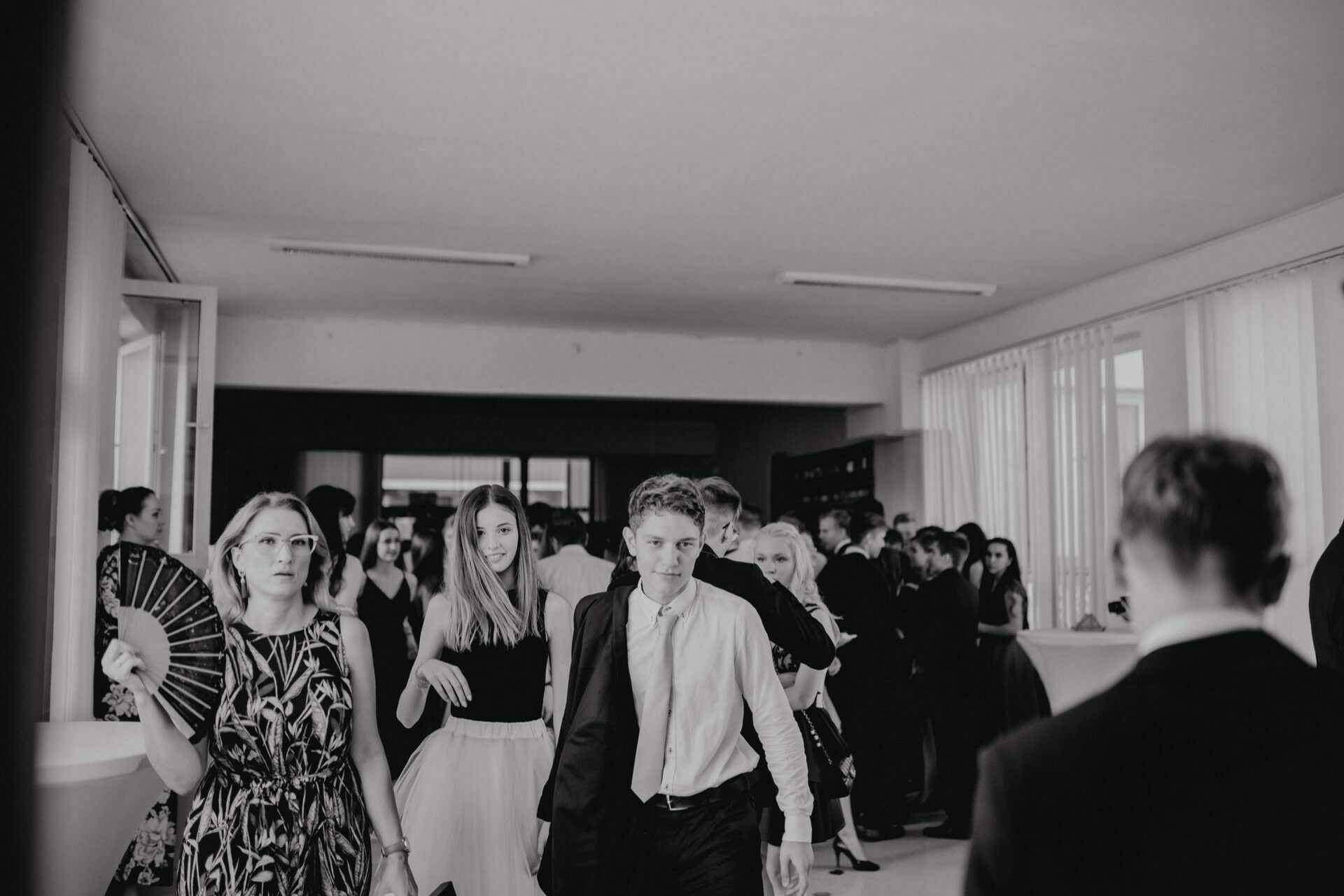 A group of people in formal attire gather in a well-lit room with large windows. Some people walk toward the camera, others meet in the background. The atmosphere seems lively and friendly. This black and white event photography, which is a photo report of the events, shows an engaging moment.   