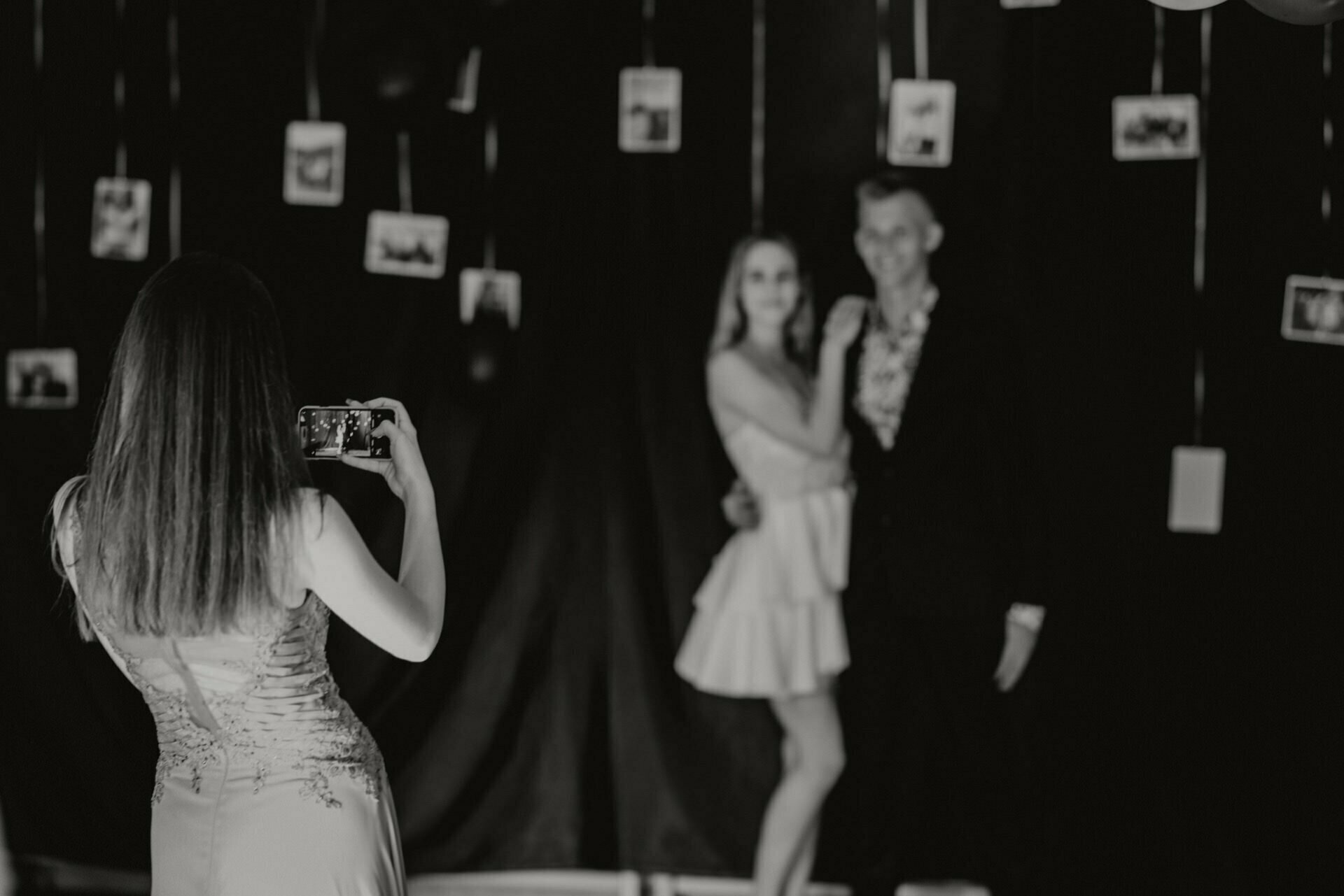 Black and white photo of a woman taking a picture of a posing couple with her phone. The couple is standing close together, with the woman on the right wearing a light-colored dress and the man on the left in a suit. In the background, pictures hanging on strings are visible, capturing this elegant event photography.  