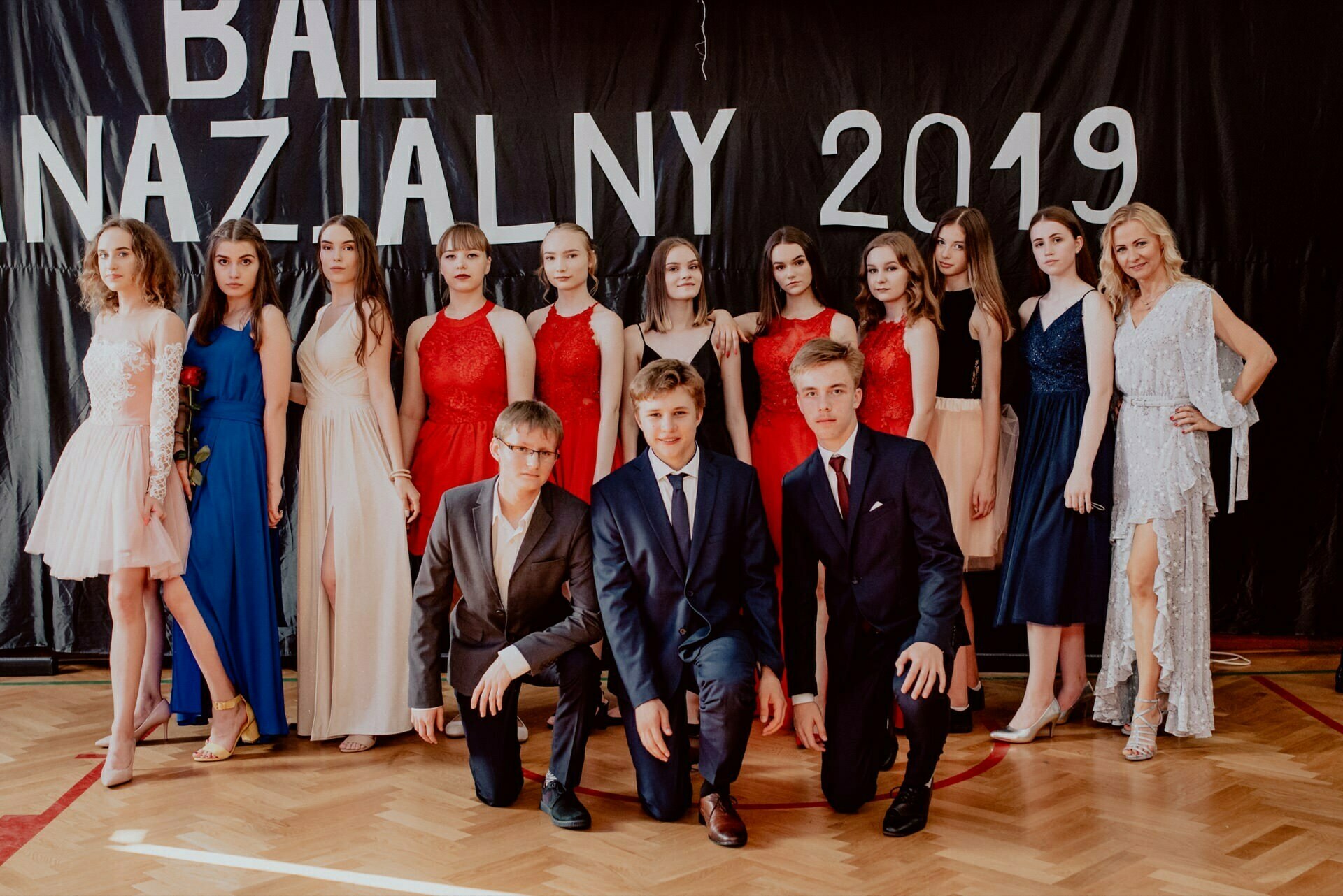 A group of well-dressed young people stand and kneel in front of a black backdrop with white letters with the words "NATIONAL BALL 2019." The girls wear elegant dresses in different colors, the boys wear suits. The floor is wooden, and they pose for a ceremonial photo taken by an event photographer Warsaw.  