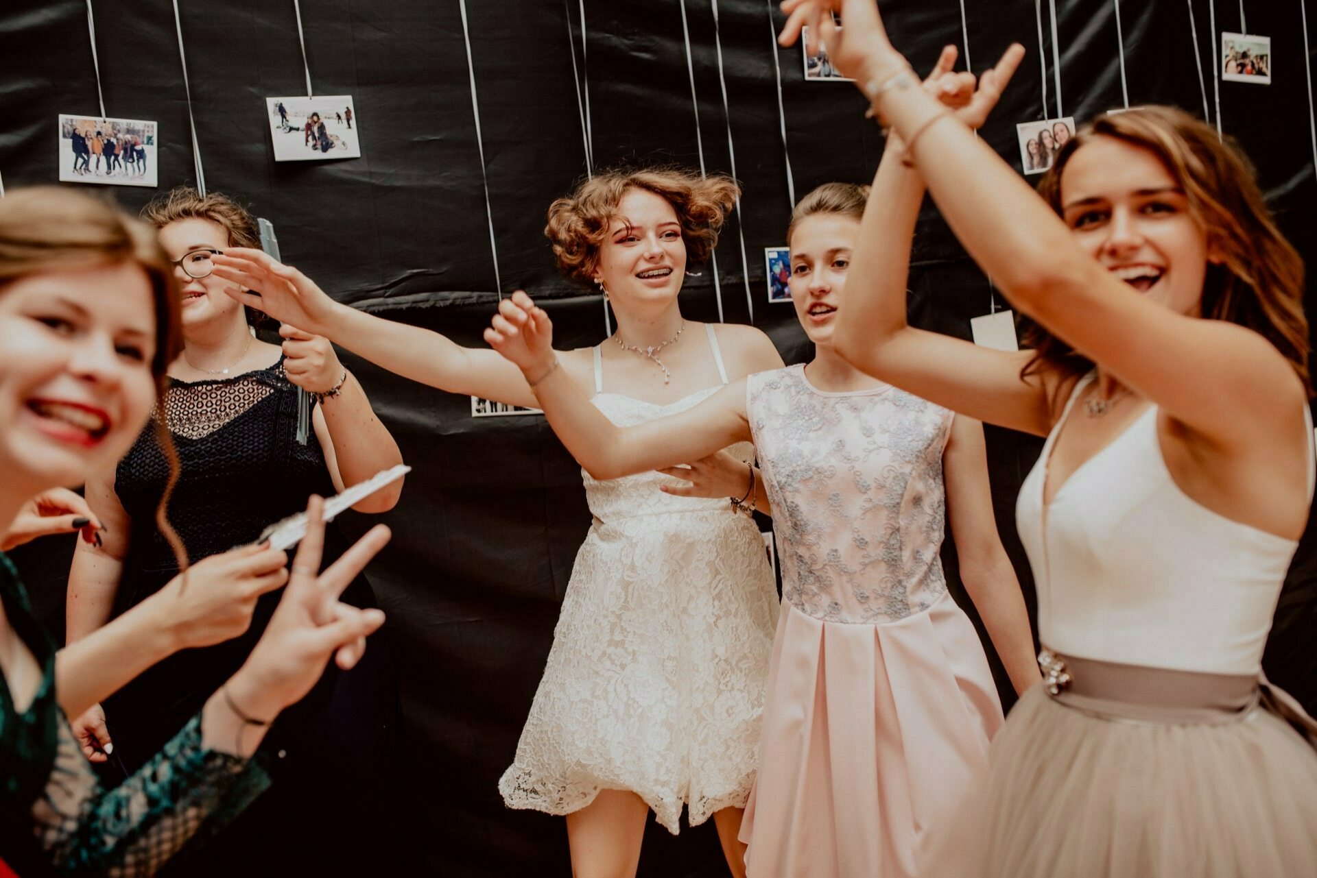 A group of young people dressed in formal attire, smiling and posing for the camera at a festive event. Some make peace signs with their hands. In the background hang photographs on strings against a black background. This lively photo report of the events perfectly captures the joyous and festive atmosphere.   