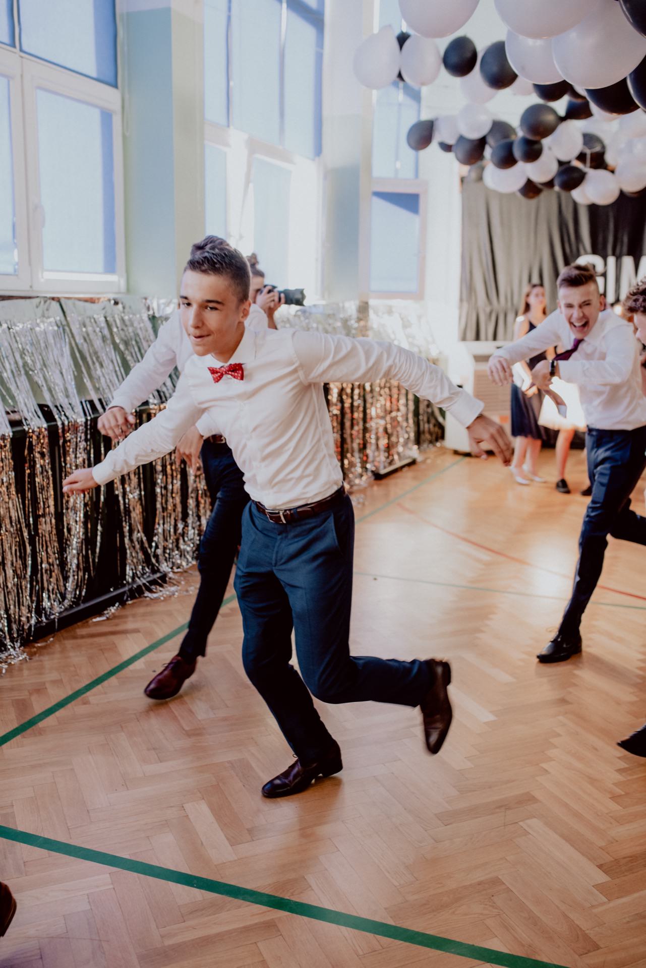 A young man in a white shirt and red bow tie dances energetically in a decorated room with black and white balloons and glittering candlelit curtains. Other people in white shirts and black pants dance in the background, creating a lively atmosphere perfect for event photography. The room has large windows that provide natural light.  