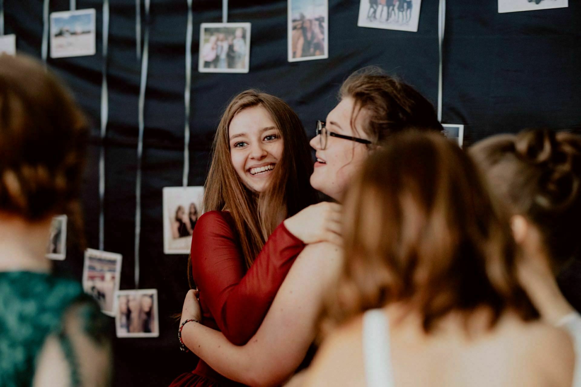 A group of women gather in a room decorated with hanging photographs. One woman, smiling radiantly, hugs another woman wearing glasses. There are strings of photos in the background, creating a nostalgic and celebratory atmosphere, perfectly captured by a skilled event photographer Warsaw event photography.  