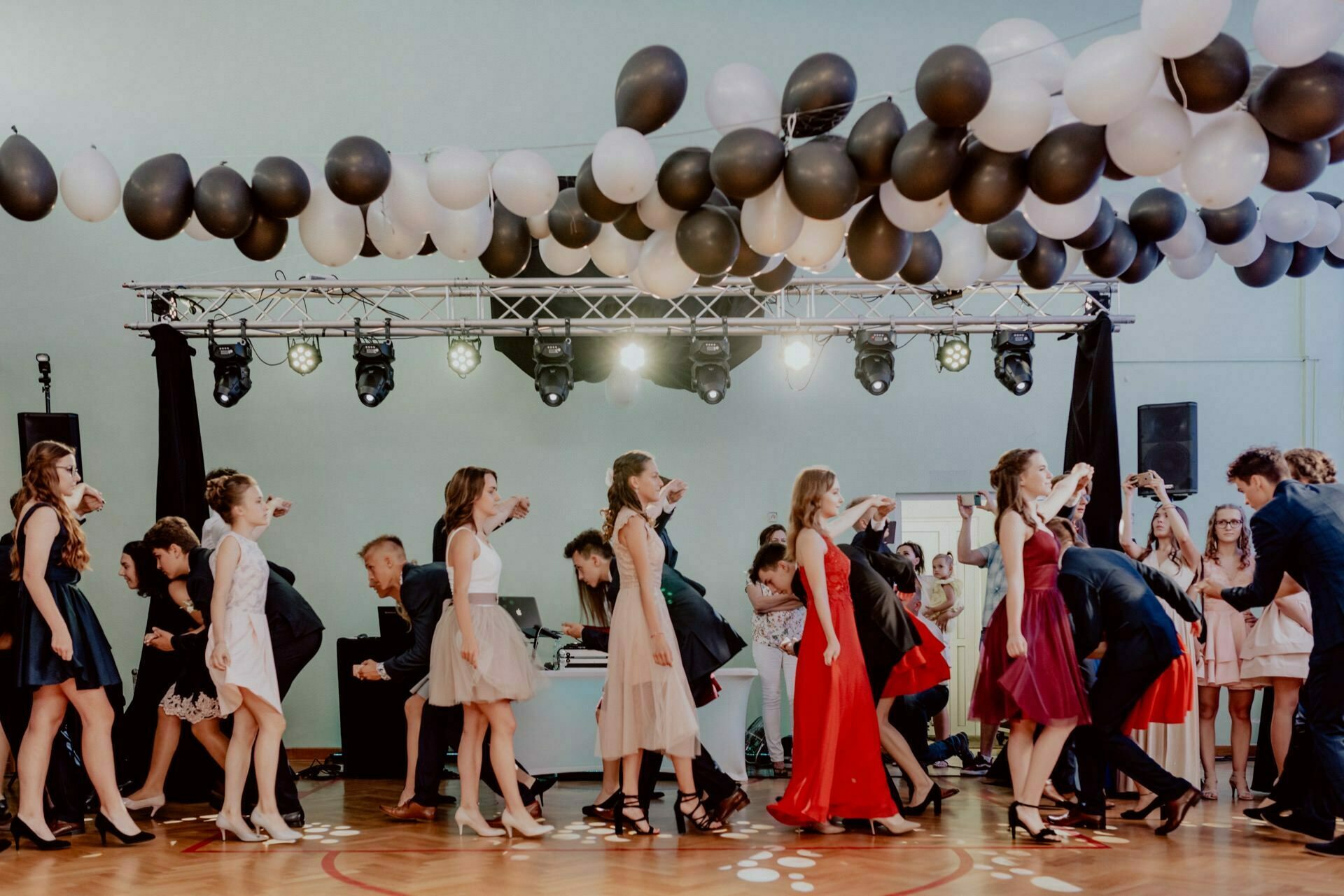 A group of people dancing in pairs in a decorated area. Black and white balloons hang from the ceiling, and stage lights illuminate the space. Many of the attendees are dressed in formal attire, including dresses and suits, perfect for photo opportunities at the event.  