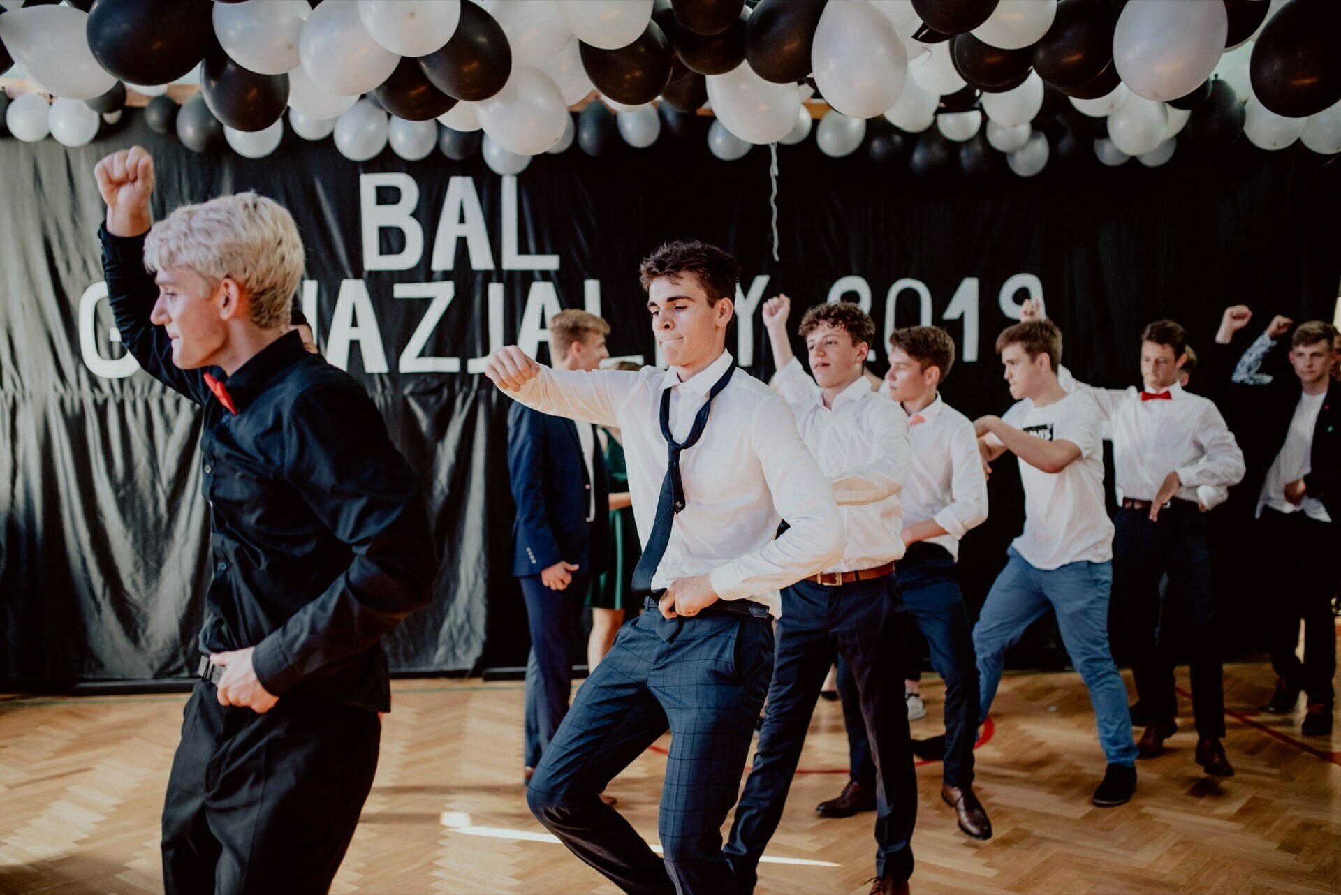 A group of young men in formal attire dance vigorously under black and white balloons. They are dressed in white shirts and dark pants, some with loose ties. This event photo report, perfectly captured by event photographer Warsaw, highlights the excitement surrounding the "2019 Gymnasium Ball.  