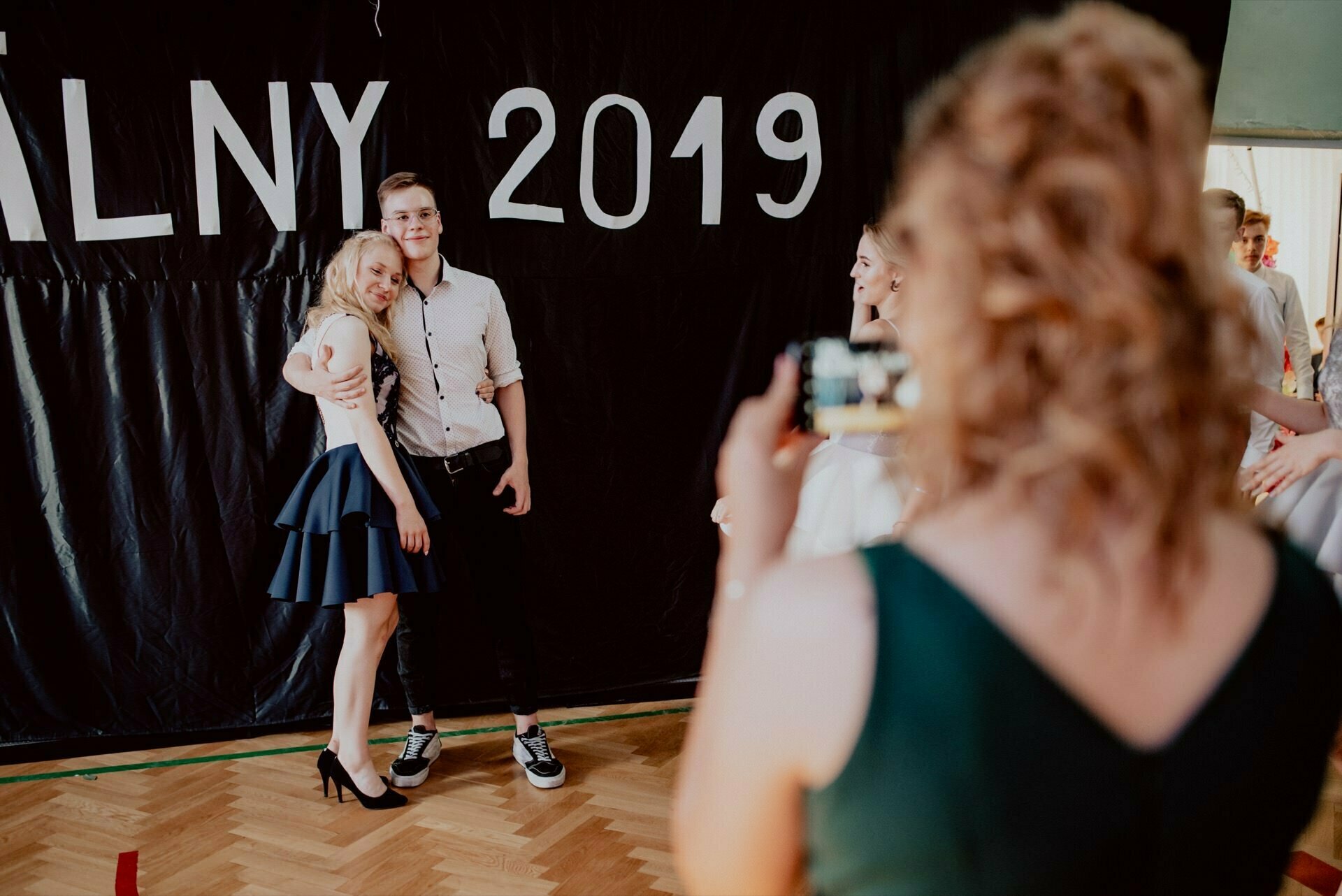 A young woman and a man stand in front of a large black banner that reads "BALNY 2019." The woman smiles and leans into the man, who puts his arm around her. A person with curly hair, dressed in a green outfit, takes a picture of them, capturing this perfect event photography moment.  