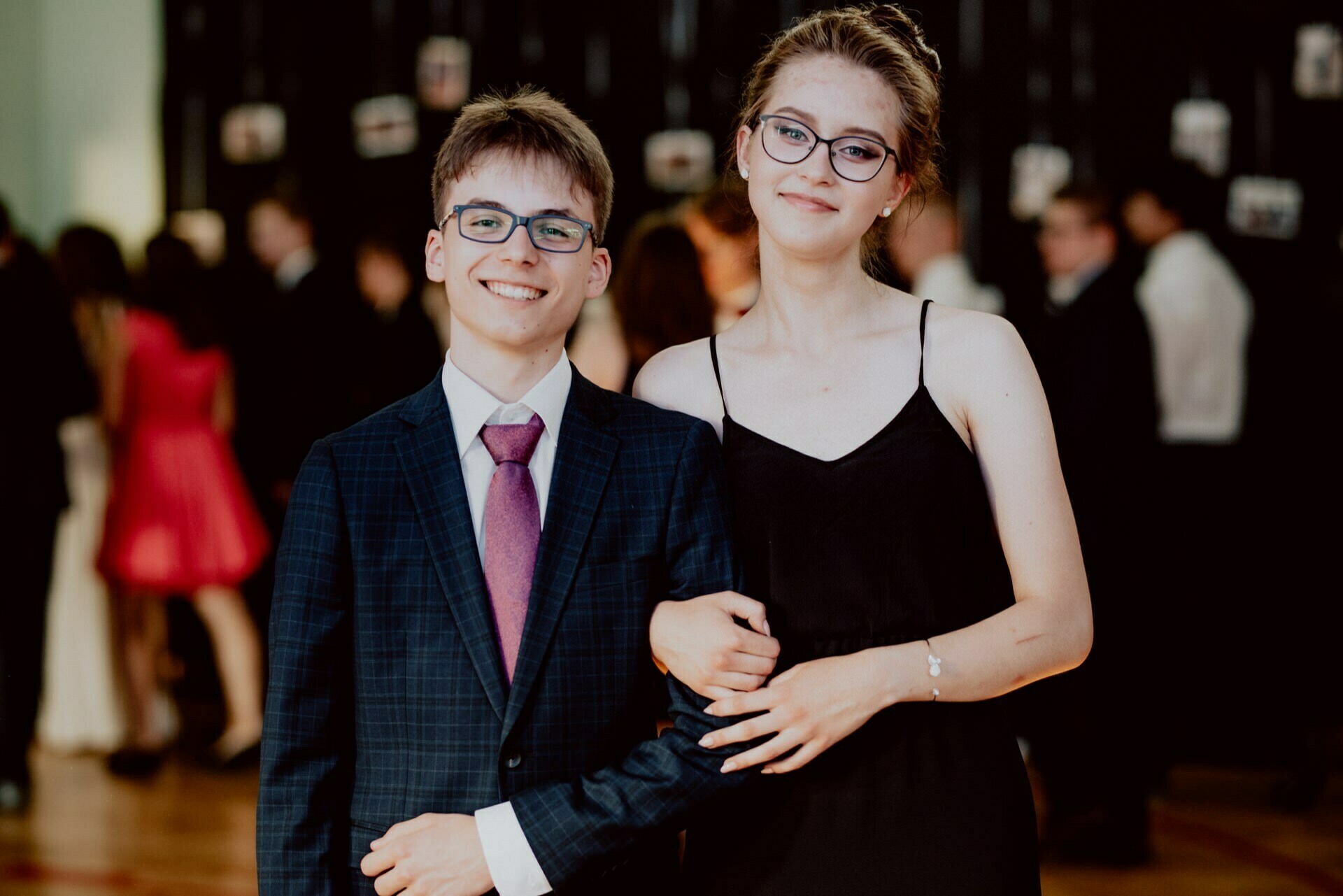 Two young people, one in a blue suit with a purple tie and glasses, and the other in a black dress and glasses, stand shoulder to shoulder and smile during a formal event. This event photography beautifully captures a festive occasion with slightly blurred people and decorations in the background. 