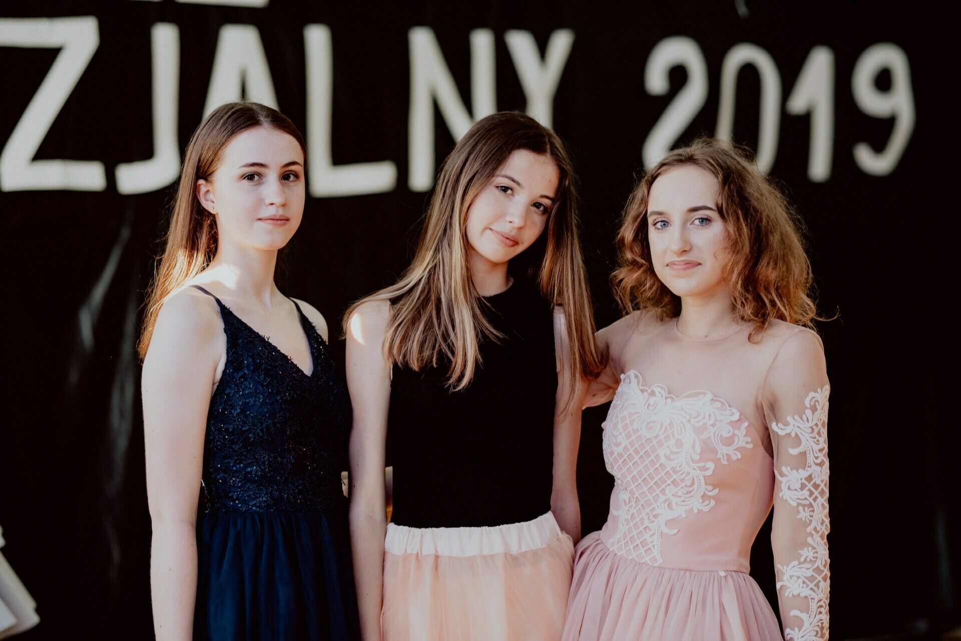 Three young women pose together, wearing elegant dresses. The one on the left is wearing a navy blue dress, the one in the middle is wearing a black top with a beige skirt, and the one on the right is wearing a pink dress with lace details. This captivating event photography captures the essence of "ZJA£NY 2019."  