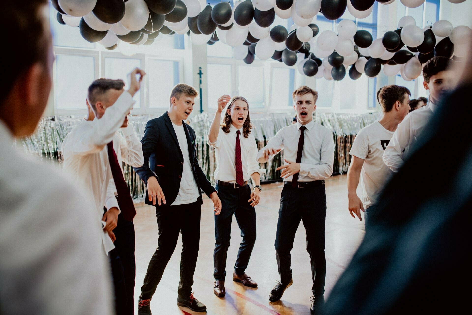 A group of teenagers in formal attire dance enthusiastically under a ceiling decorated with black and white balloons. Some wear ties and jackets, while others wear white shirts. The event is reminiscent of a formal school dance, as beautifully captured by an event photo by event photographer warsaw.  