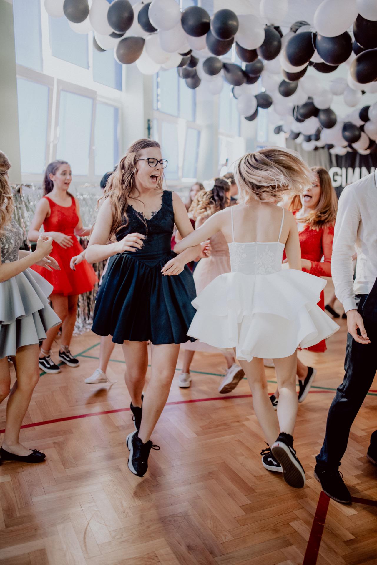 A group of people dance in a decorated room with black and white balloons on the ceiling. Two women in the foreground twirl, one in a black dress and the other in a white dress. Both seem to be having a good time on the wooden floor. It's the perfect scene for any *event photographer warsaw*, capturing sincere joy.   