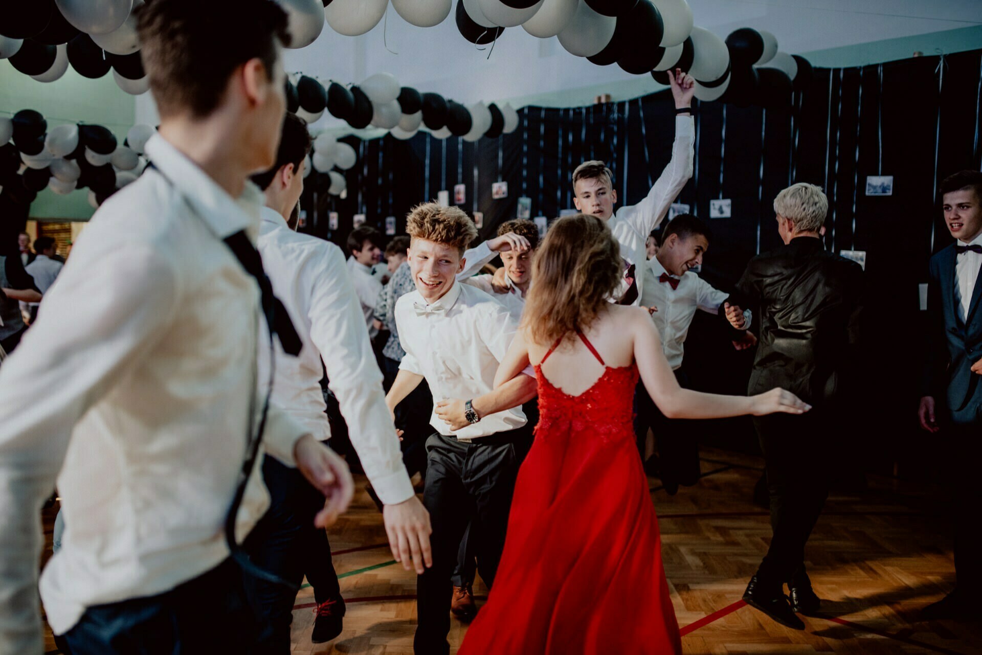 A group of formally dressed people are dancing and having fun under black and white balloons. One woman in a red dress is in the foreground, while others smile and move energetically around her during what appears to be a formal event or celebration - perfect for event photography. 