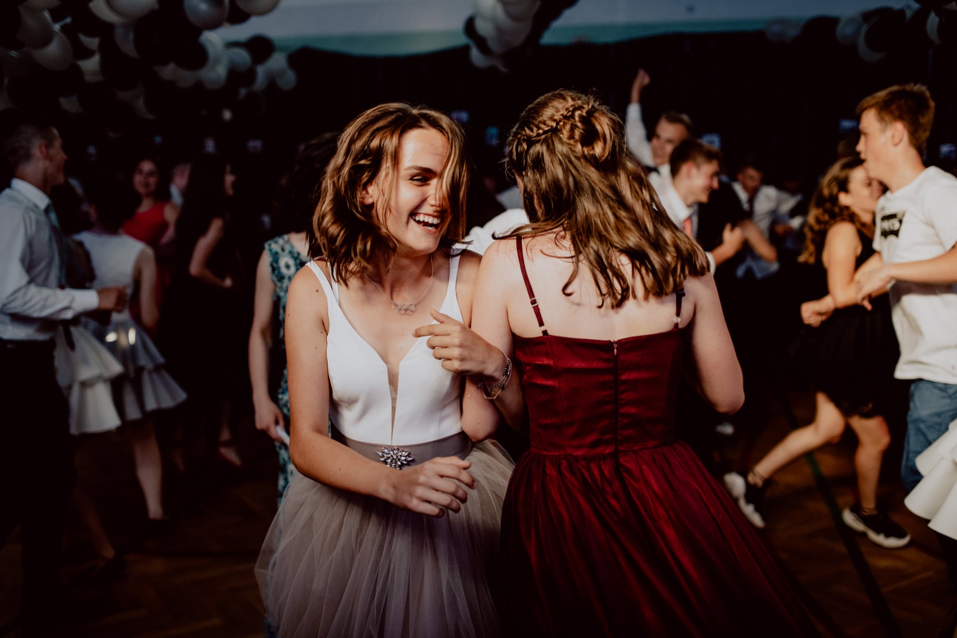 A lively party scene with people dancing and socializing. In the foreground, two women in dresses are laughing and having fun. One is dressed in a white top and gray skirt, and the other in a red dress. The background captured with event photography is filled with other partygoers.   