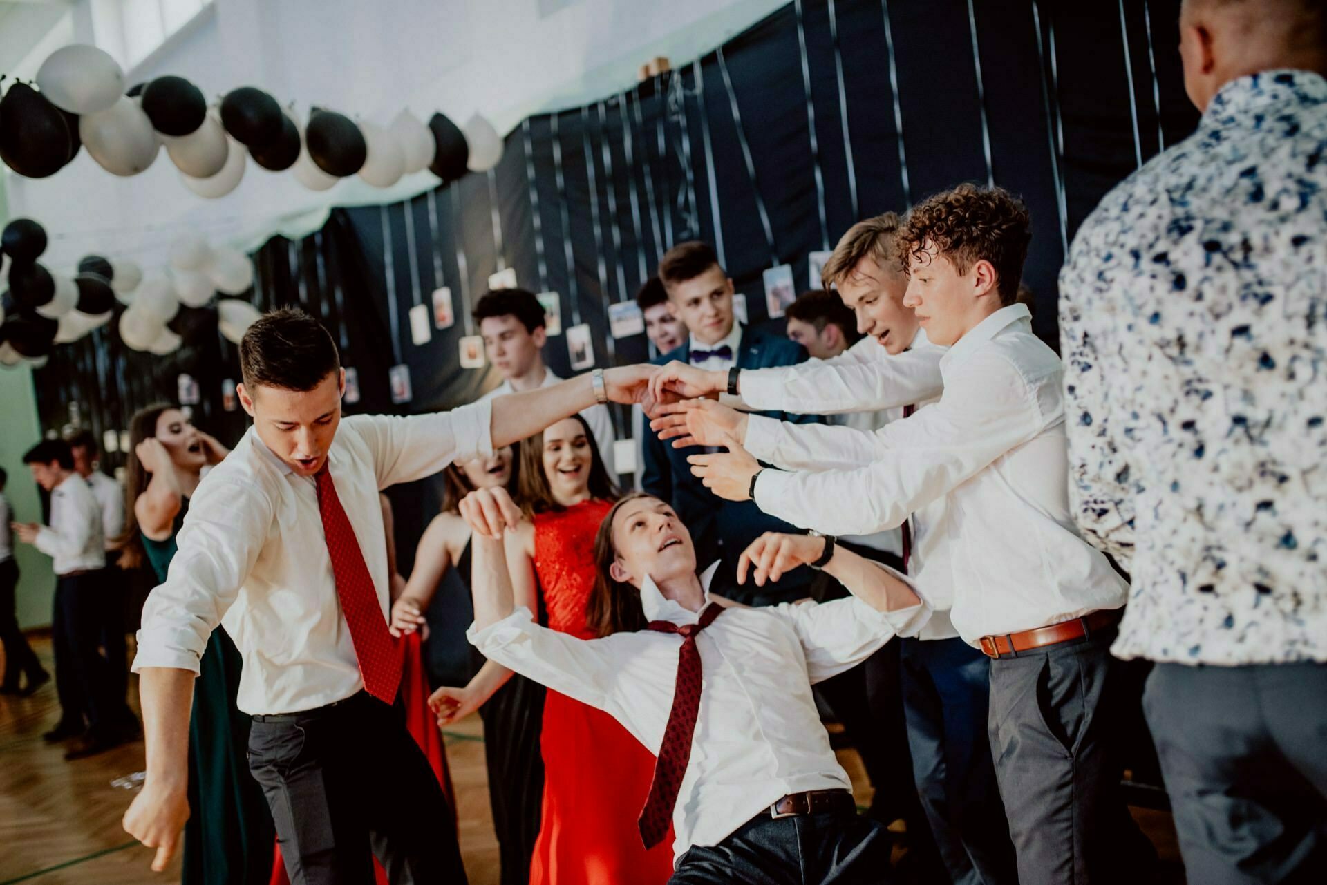 A group of people dressed in formal attire dance energetically in a decorated room with balloons and hanging lights. One person is playfully lifted up while others around her laugh and enjoy the moment, perfectly captured as part of a wonderful photo report of the events. 