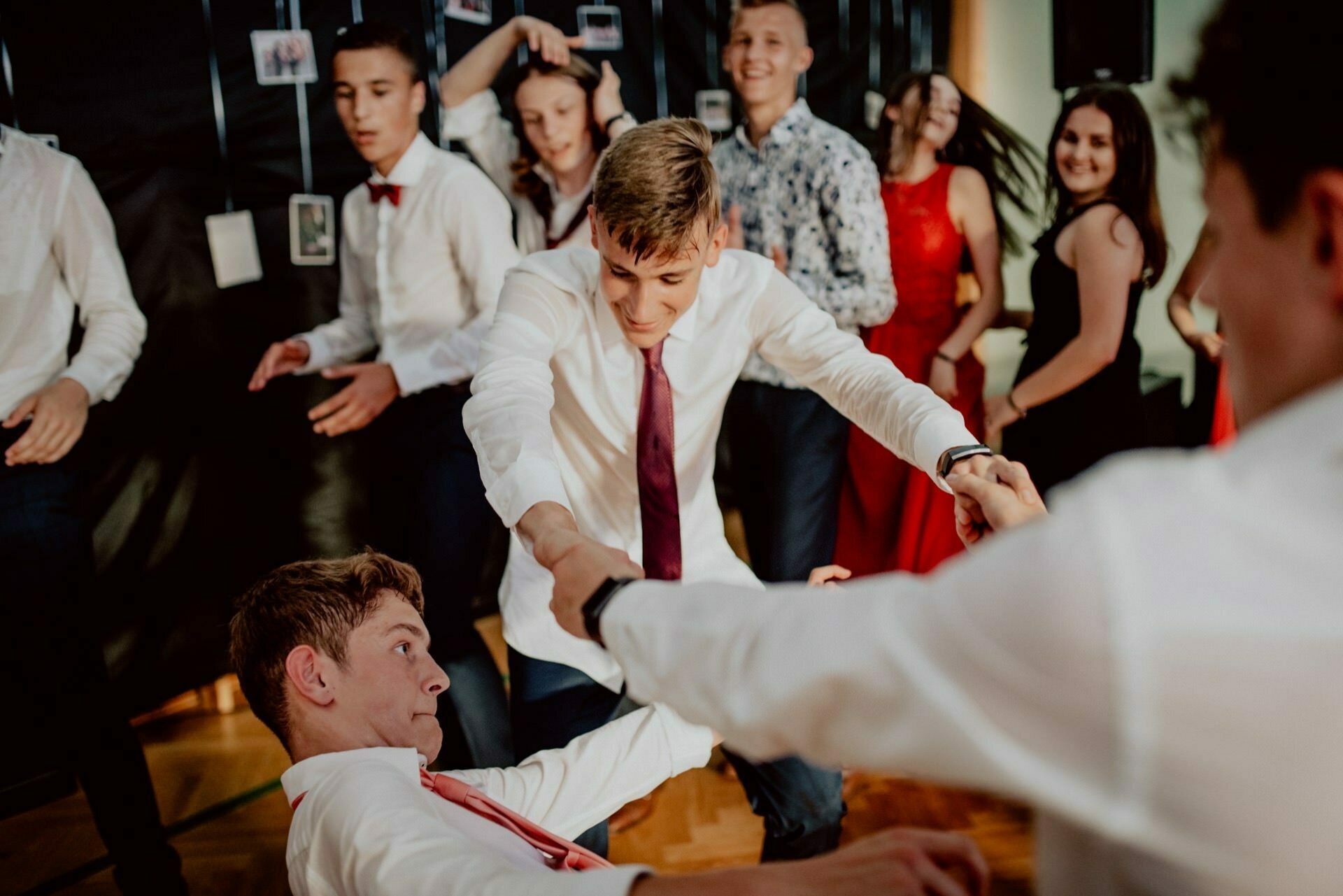 A group of teenagers dance at a lively party. A teenager in a white shirt and red tie helps another get up from the floor, holding his hand. Others around them are smiling and enjoying the lively atmosphere, perfect for photo coverage of the events. In the background, photos hang on strings.   