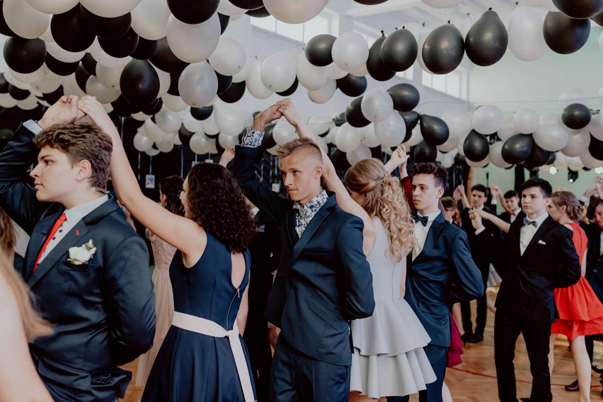 People dressed in formal attire dance in pairs at a party under an array of black and white balloons. The venue has high ceilings and the atmosphere seems festive, with attendees dancing in ballroom style. A skilled event photographer will capture the elegance of this unforgettable evening.  