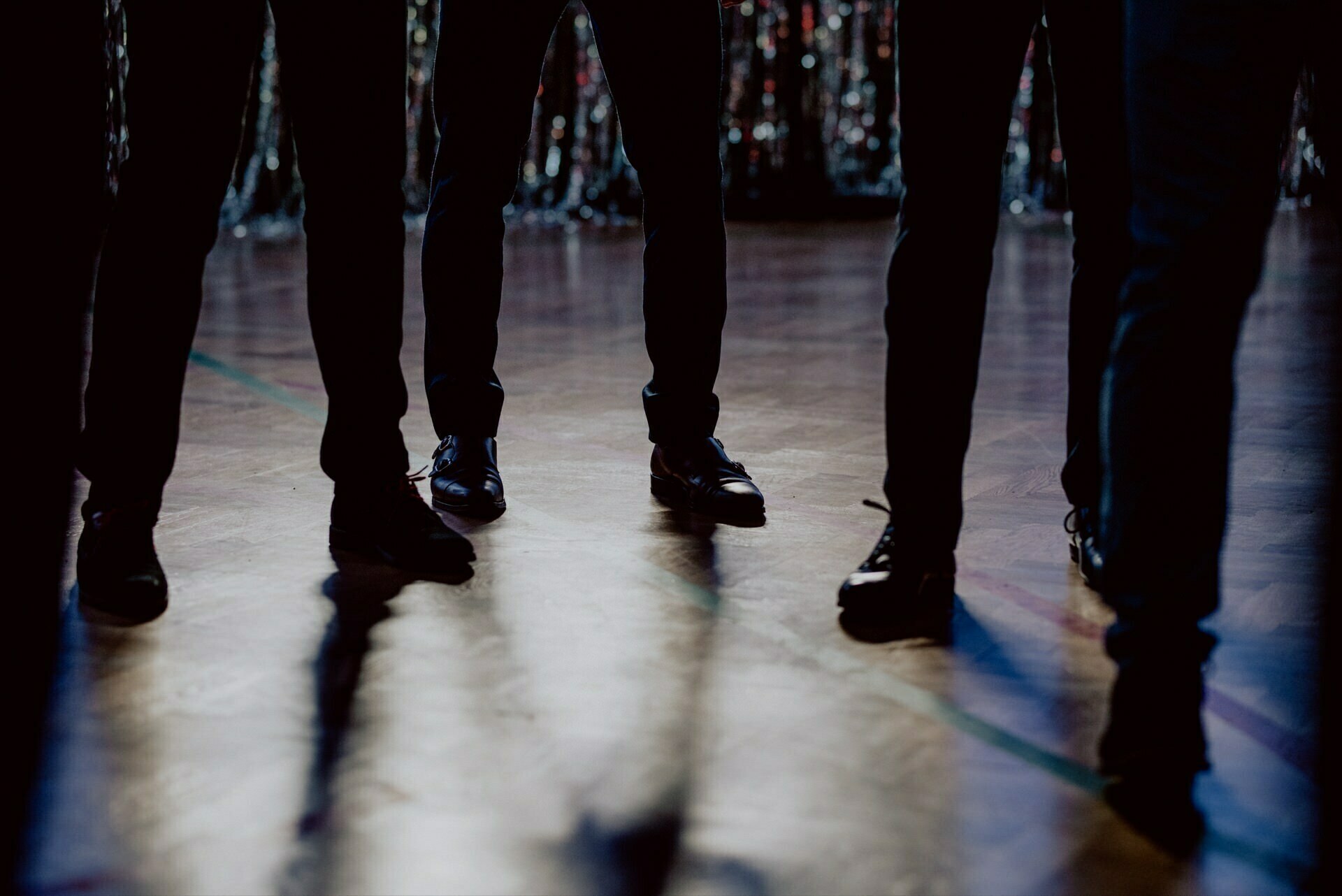 Silhouette showing the legs and feet of four people dressed in dark pants and shoes, standing on a polished floor. The background is blurred by indistinct lights, suggesting an indoor setting. Shadows of their feet are cast on the floor, creating a dramatic scene ideal for event photography.  