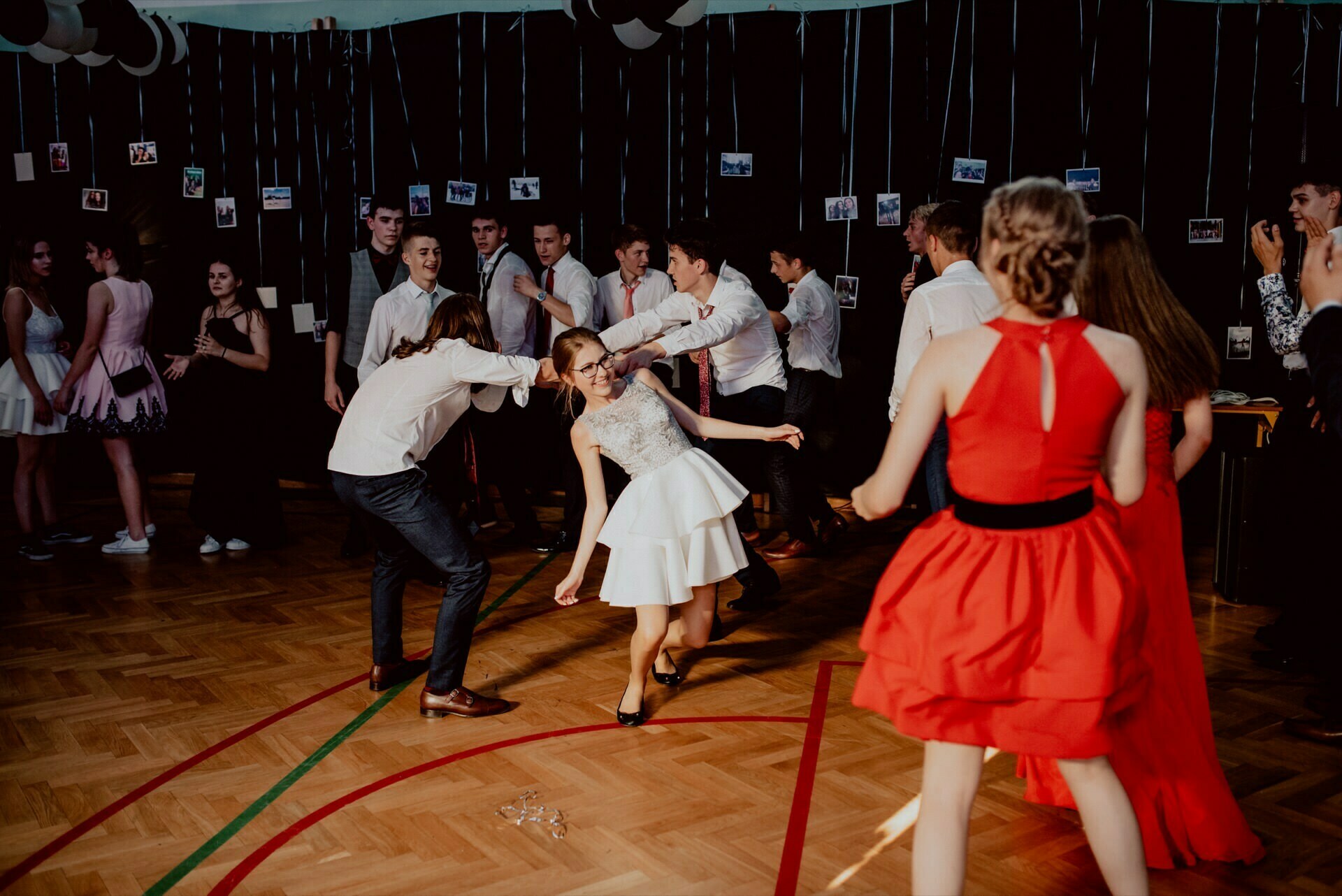 A group of young people dance vigorously in the decorated hall, capturing the essence of the photo-report of the events. Most are dressed in formal or semi-formal attire, the girls in dresses, the boys in shirts and pants. In the background, hanging photos and streamers create a lively and joyful atmosphere.  