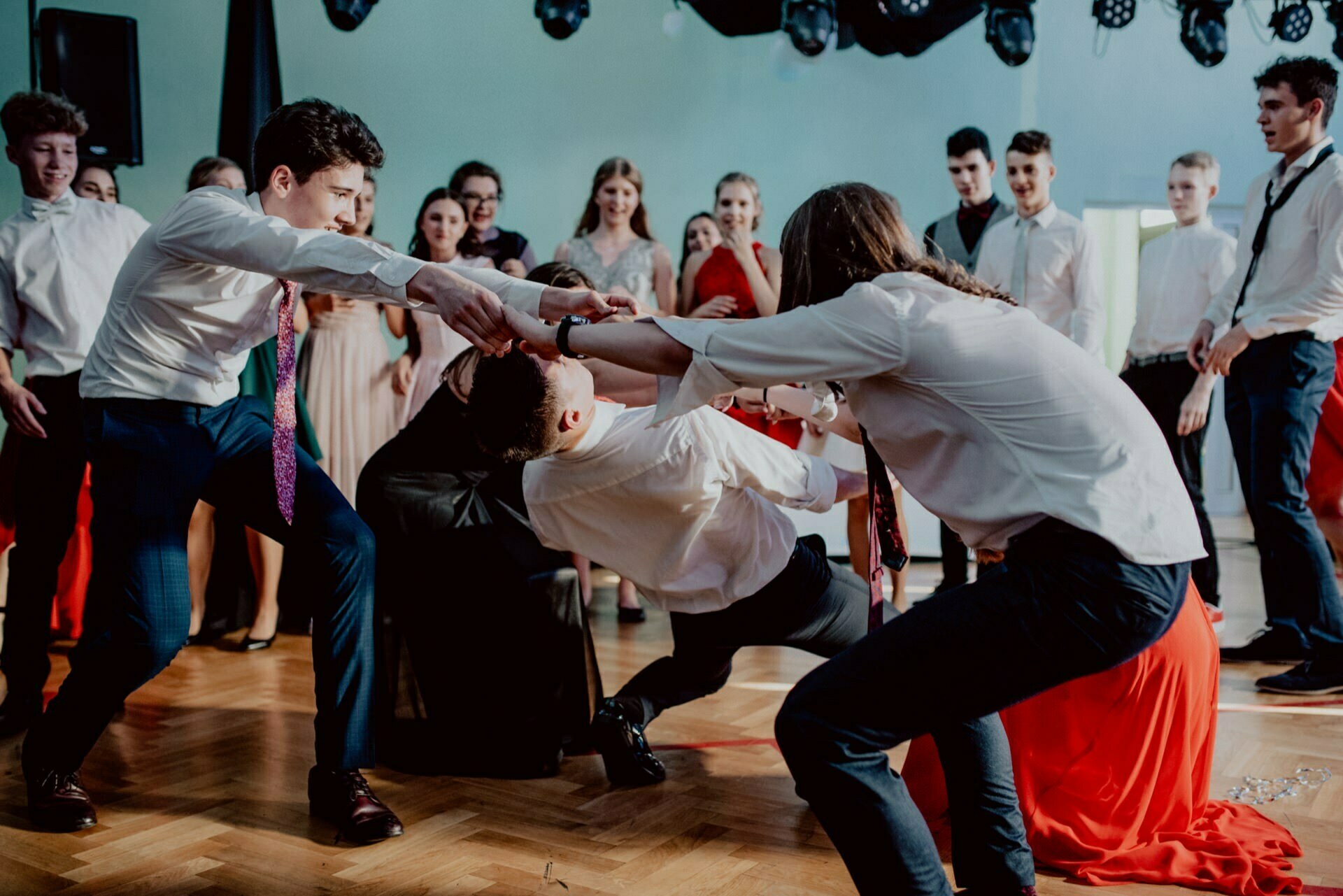 A group of people are in a well-lit room, dressed formally. Two men playfully pull another man, who leans back and almost falls. Other people in the background are watching and laughing, creating a lively and festive atmosphere that is perfectly captured by the photo report of the events.  