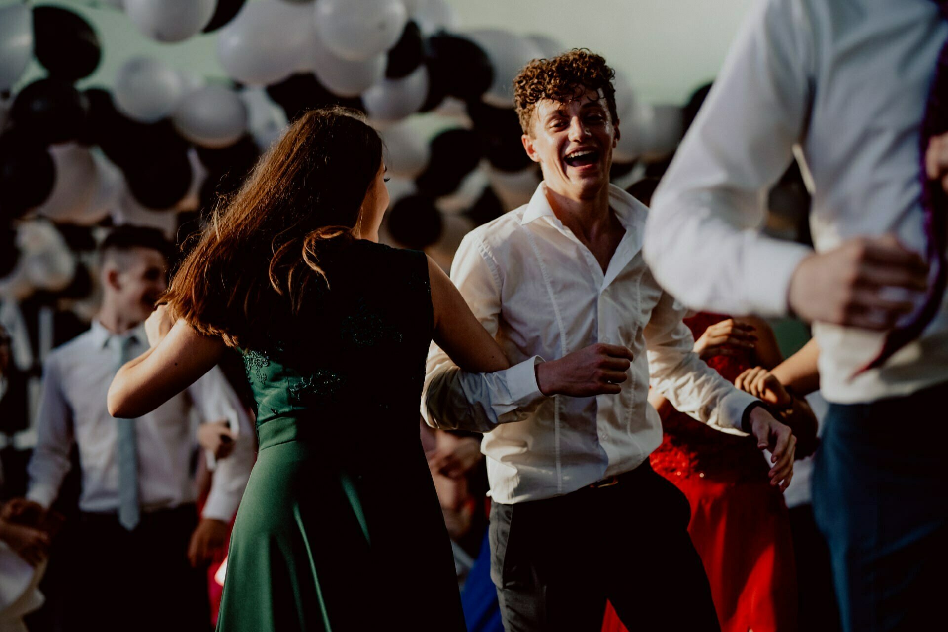 A group of people dancing in a festively decorated room with black and white balloons. In the center, a smiling person in a white shirt dances with another person dressed in a green dress. The atmosphere seems joyful and lively, perfectly captured by the event photographer Warsaw, creating an unforgettable photo report of the events.  