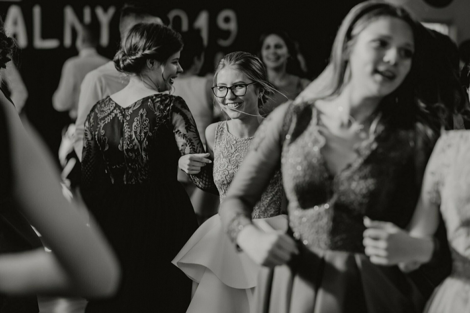 A group of young people in formal attire joyfully dance at a party. The scene was captured by a black and white event photographer Warsaw, focusing on a smiling woman in glasses and a detailed dress. The caption in the background reads "2019." This event photography captures the moment beautifully.   