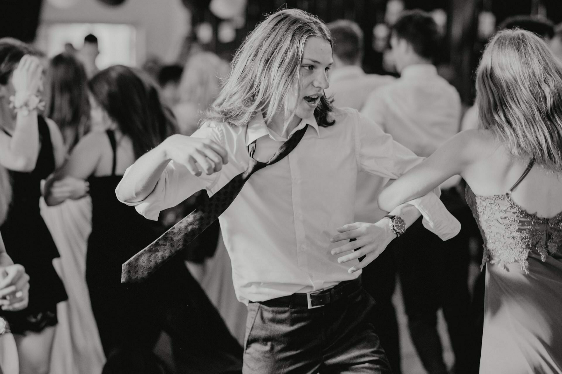 A young man with long hair, wearing a white shirt and dark tie, is dancing enthusiastically at a crowded party. He has an animated expression and lively posture, while others around him also seem to be dancing. This black-and-white event photo captures the lively energy of the event.  