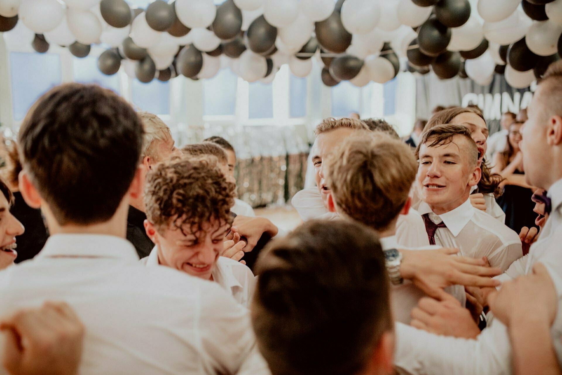 A group of young people dressed in formal attire joyfully huddle under an arrangement of black and white balloons. This scene seems to come from a festive indoor event with everyone smiling and laughing - truly a moment perfectly captured in event photography. 