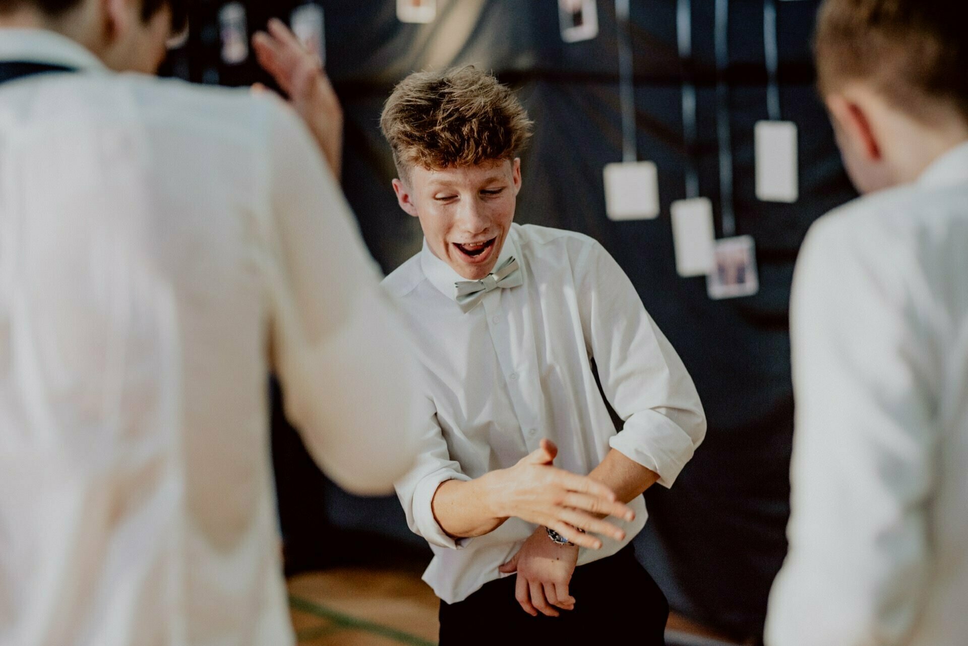 A young man dressed in a white shirt and light blue bow tie dances energetically, surrounded by other people in similar attire. He moves his arms enthusiastically. A dimly lit backdrop, decorated with hanging photographs, sums up the lively photo recounting of events.  