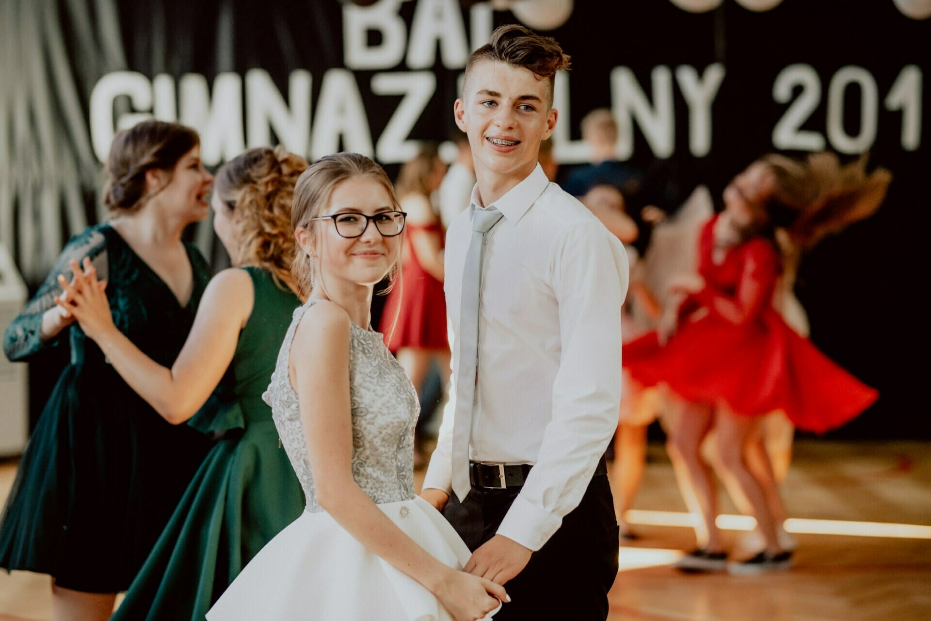 A young couple dances at a school dance, smiling for the camera. The young woman is wearing glasses and a dress, while the young man is wearing a white shirt and tie. There are other couples dancing in the background, and the sign on the wall reads "Junior High School 2019." This captivating image perfectly captures the essence of event photography.   