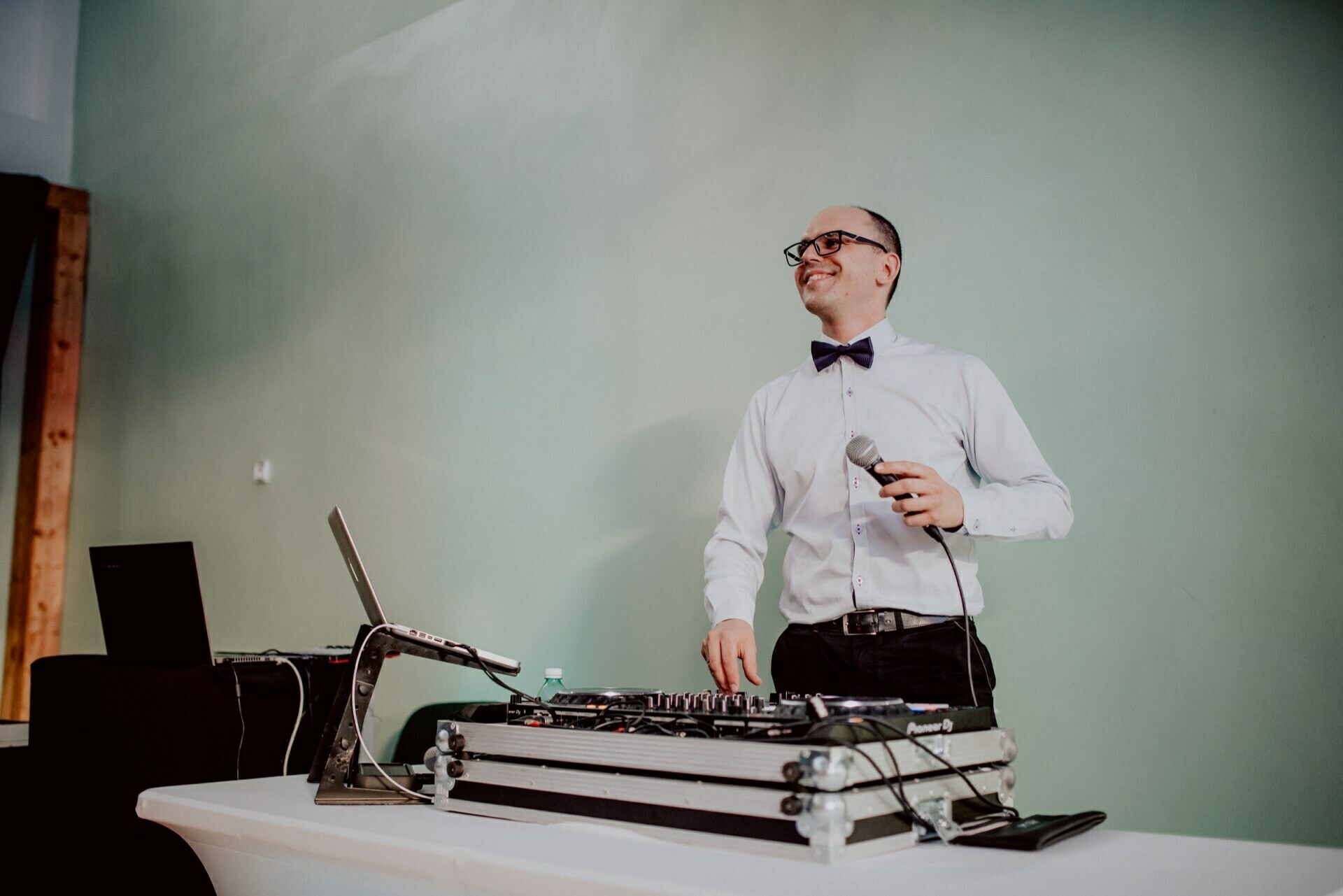 A man in a white shirt and black bow tie stands behind a table with DJ equipment, holding a microphone and smiling. He gives the impression of a DJ at a party, perfectly capturing the spirit often found in event photography. The background is a bright green wall, which adds to the vibrant atmosphere.  