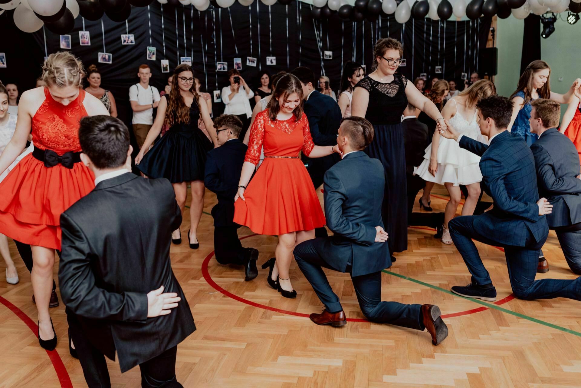 A group of people dressed in formal attire dance in pairs during a party held in a room decorated with black and white balloons. Some women in red dresses and men in suits perform choreographed dance moves while the crowd looks on in the background. This captures a vibrant photo-op of the events.  