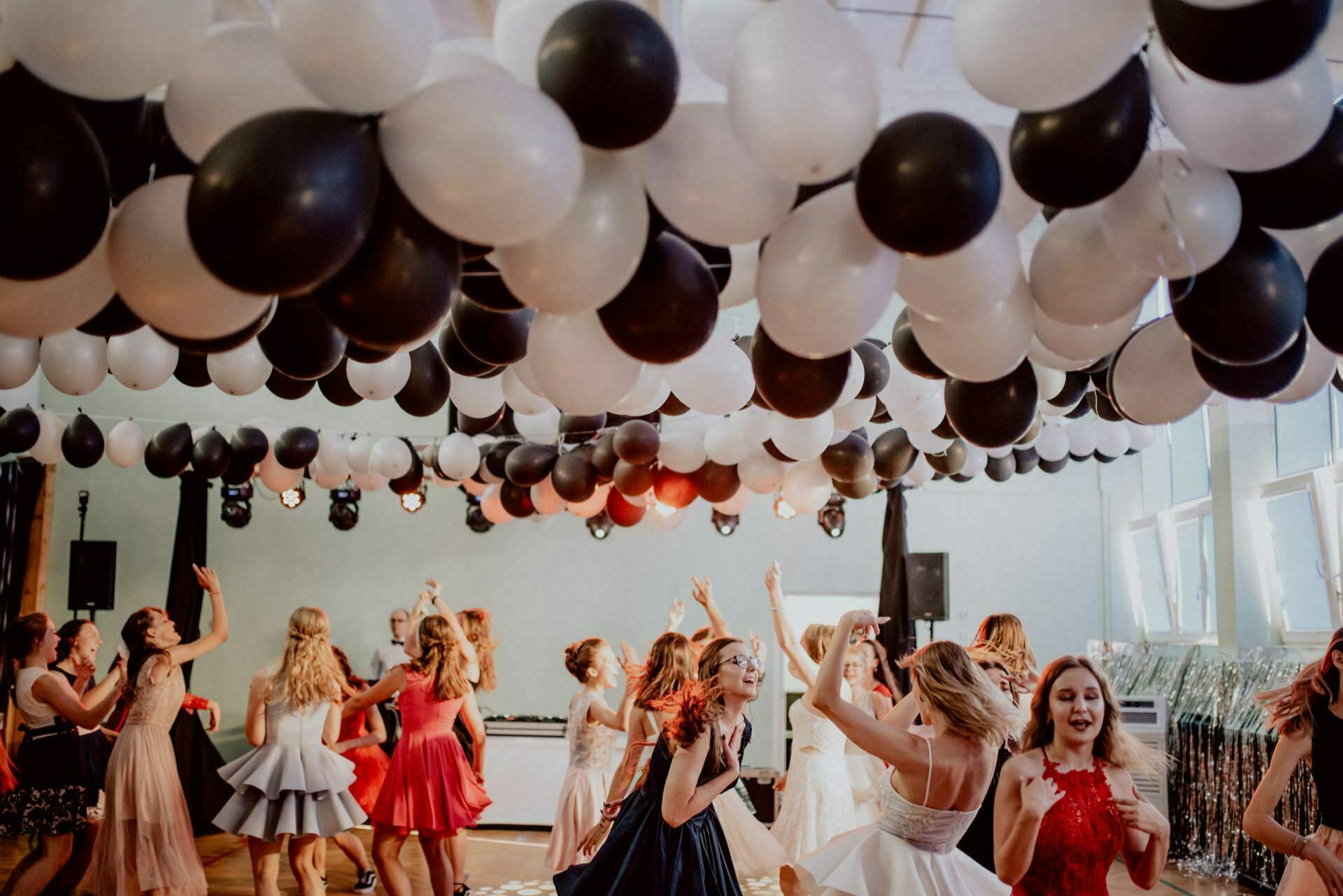 A lively party with people dancing under a ceiling filled with black and white balloons. Many of the attendees are wearing formal dresses, and the room is brightly lit with large windows on one side. The atmosphere seems joyful and festive, perfect for an event photographer Warsaw to capture stunning photos of the event.  