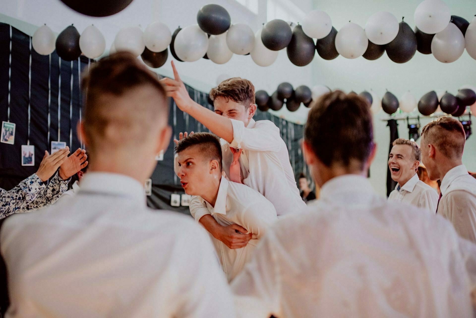 A group of young men dressed in white shirts joyfully interact at a party held in a room decorated with black and white balloons. One man lies on the back of another, pointing and smiling, while the others laugh and chat with each other in a lively atmosphere. Ideal for event photo coverage by any qualified event photographer warsaw.  