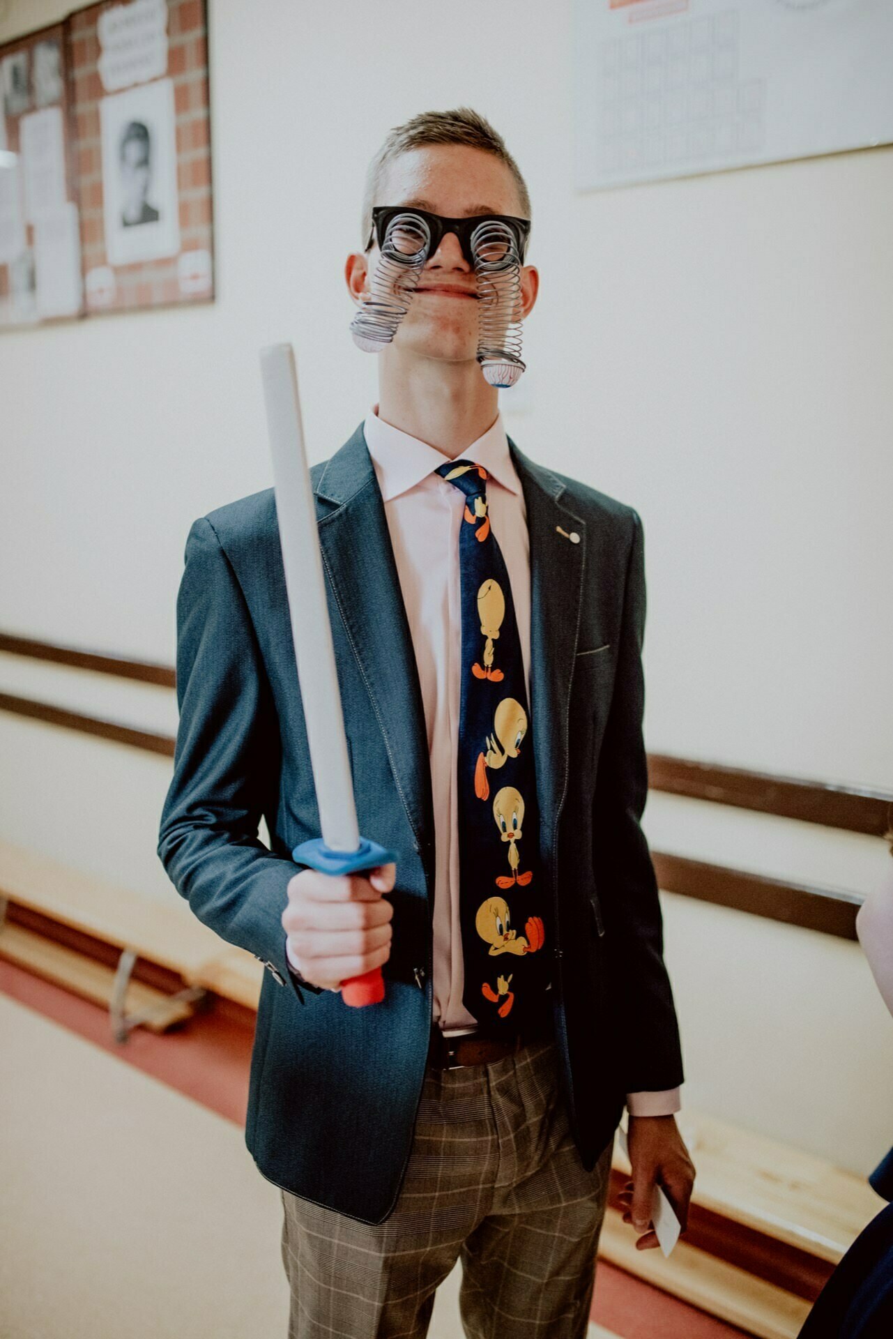 A person in a suit and tie stands in a room, holding a toy sword. Their funny glasses mimic swirling eyes, and the tie has a cartoon character design. In the background are wooden benches, bulletin boards and a substitute for a periodic table - a perfect scene for event photography by event photographer Warsaw.  