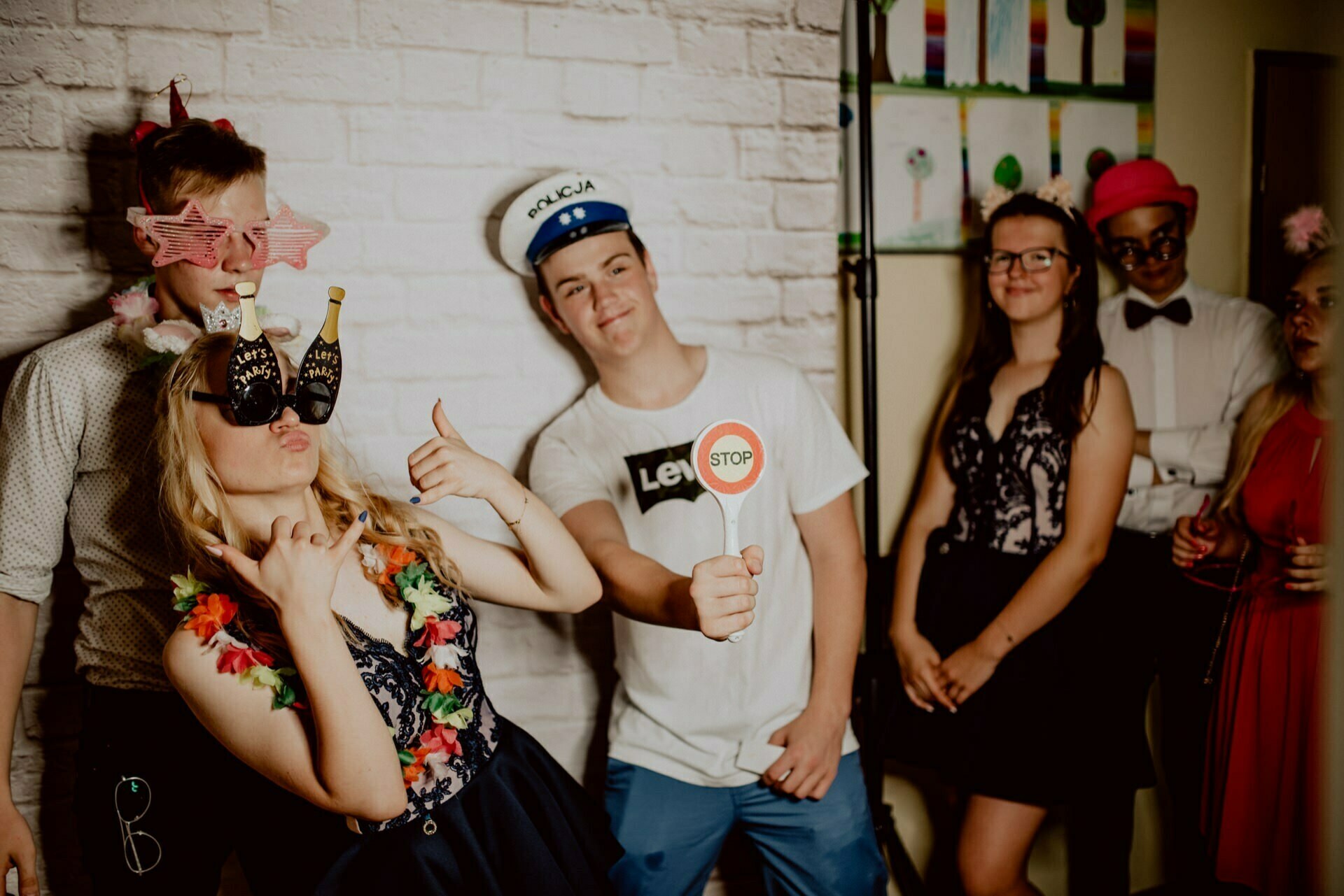 A group of six young people dressed in party attire playfully pose for a photo, perfect for event photojournalism. One person is wearing large sunglasses, another is holding a "Stop" sign, and others are wearing various party accessories such as hats and glasses. They are standing against a white brick wall.  