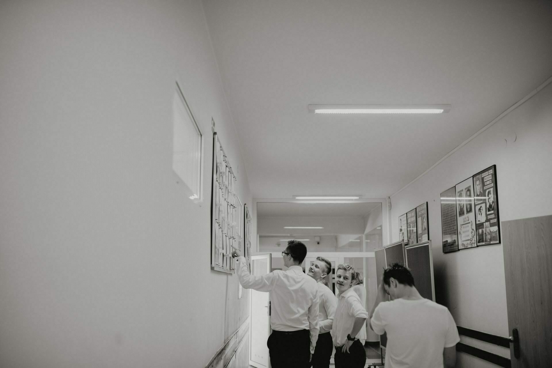 A group of four people dressed in formal attire stand in a brightly lit corridor and look at a bulletin board. The scene, captured in black and white, depicts framed pictures hanging on the walls. This photo-reportage of events perfectly captures the atmosphere and elegance of the moment.  