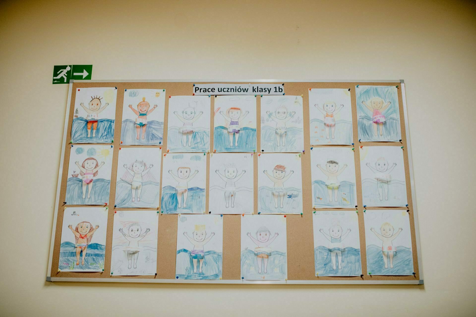 The bulletin board displays children's drawings, each depicting a human figure with hands raised, against a blue background. At the top of the board, the caption reads "Class 1b students' work," which means "Class 1b students' work." It is reminiscent of an amusing photo-report of events.  