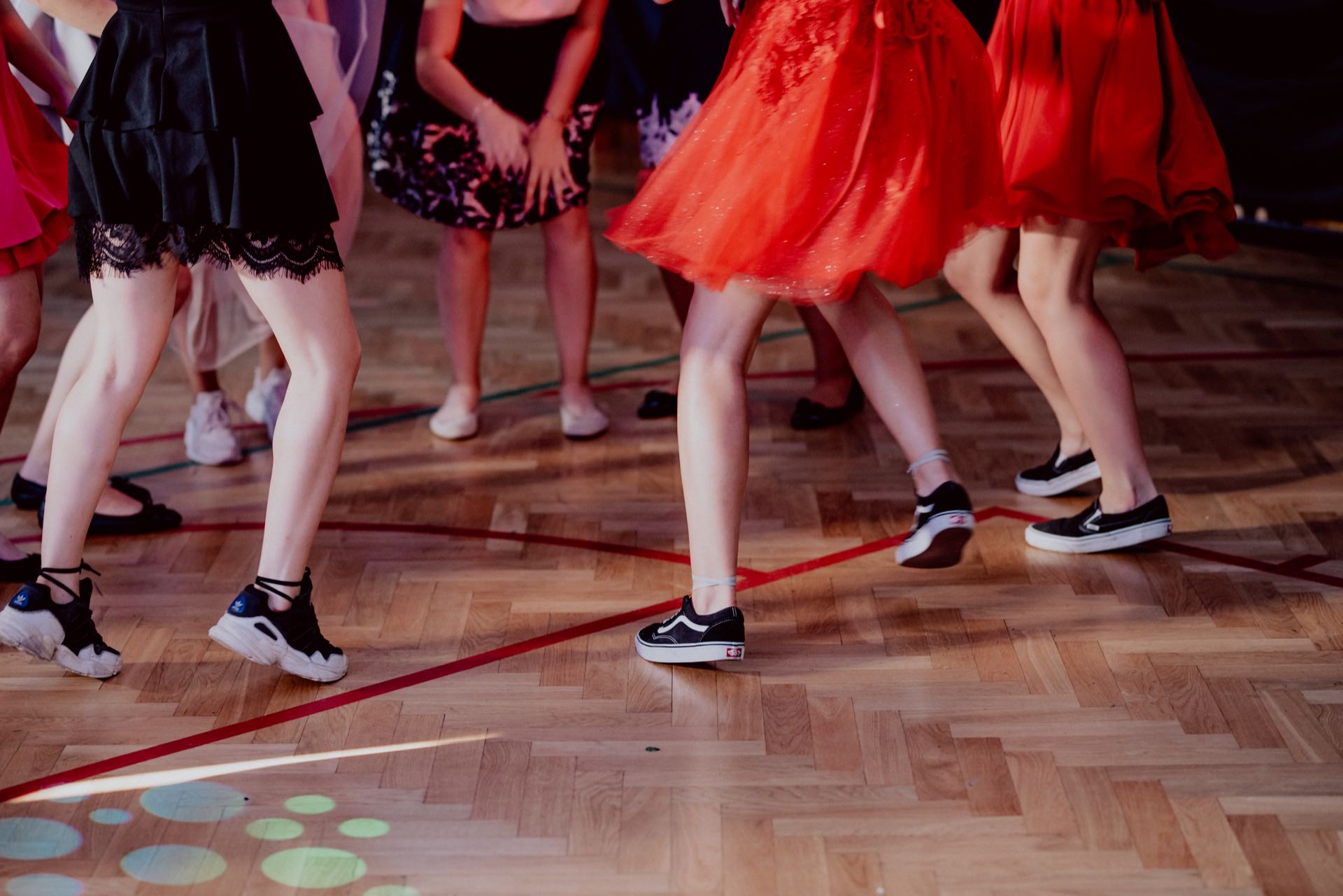 A group of people dressed in various dresses and everyday shoes dance on a wooden floor. Their faces are not visible, capturing the movement and energy of the dance. Among the styles, red dresses and sneakers dominate, perfectly capturing the photo-relation of the events with spontaneity.  