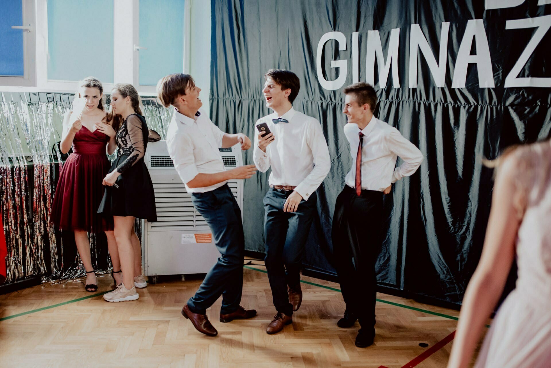 A group of young men in formal attire are laughing and sharing a phone against a black background with the word "GIMNAZ," beautifully captured in an event photo. In the background, several young women in dresses and one in tennis shoes are chatting by a wall decorated with silver streamers. 