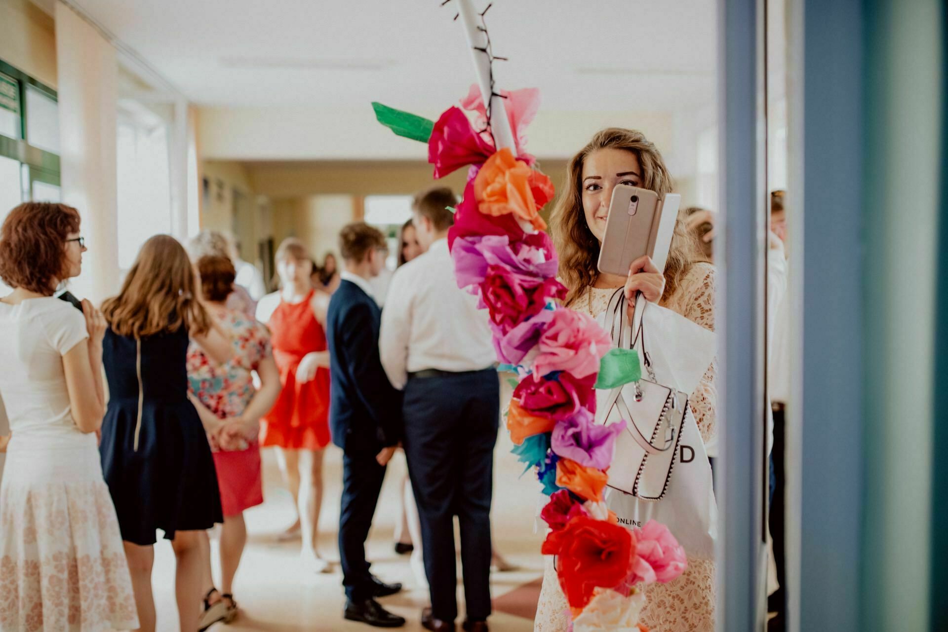 A young woman holds a smartphone and colorful paper flowers in the hallway, smiling at the camera. In the background, a group of people dressed in semi-formal clothes stand and talk. The scene seems bright and cheerful, perfectly capturing the essence of the photo report of the events.  