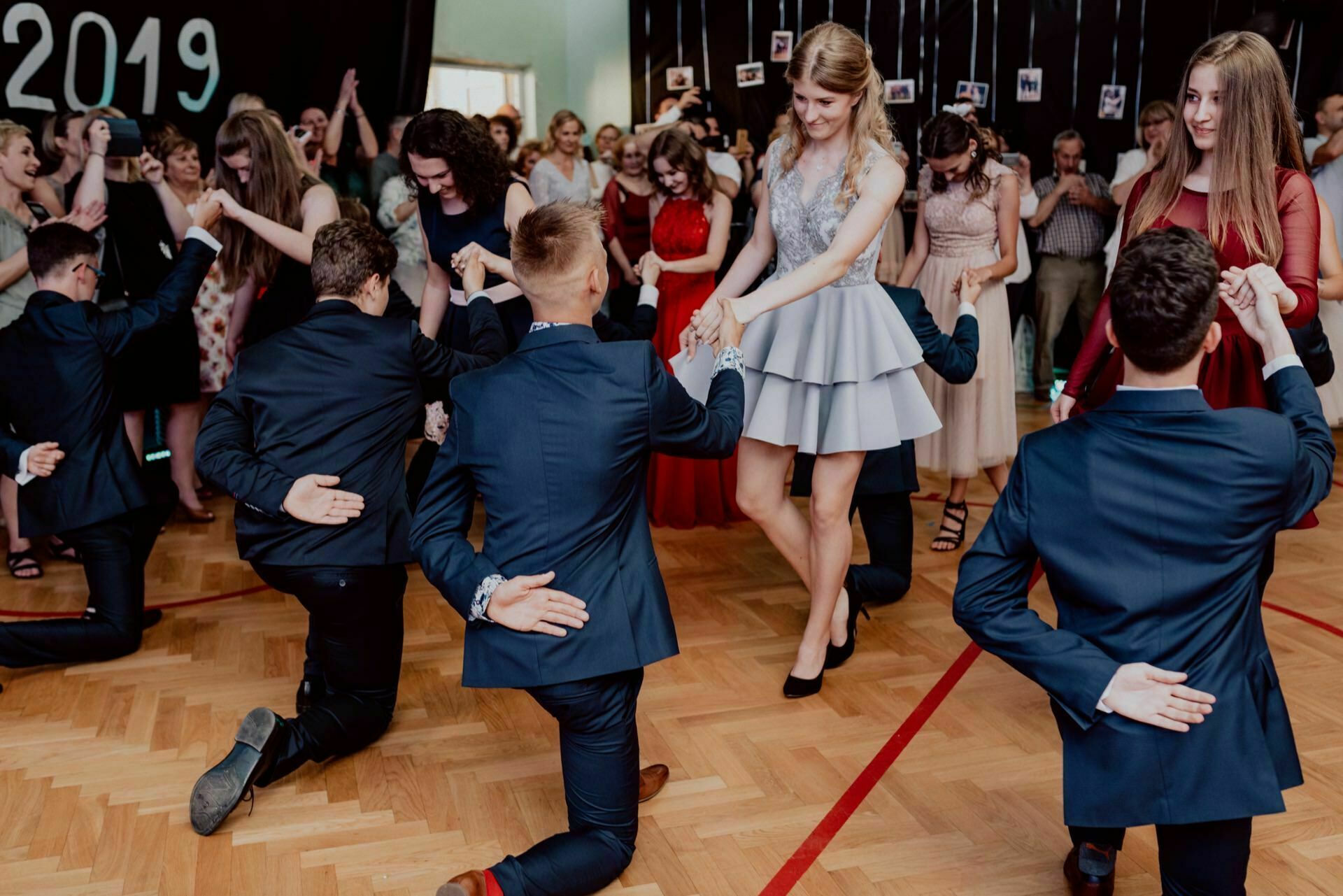 The dance involves a group of young adults dressed in formal attire. Several men kneel, holding hands with women in elegant dresses. Spectators in the background are watching and taking pictures, capturing a beautiful photo report of the events. On the left side of the image, a large inscription "2019" is visible.   